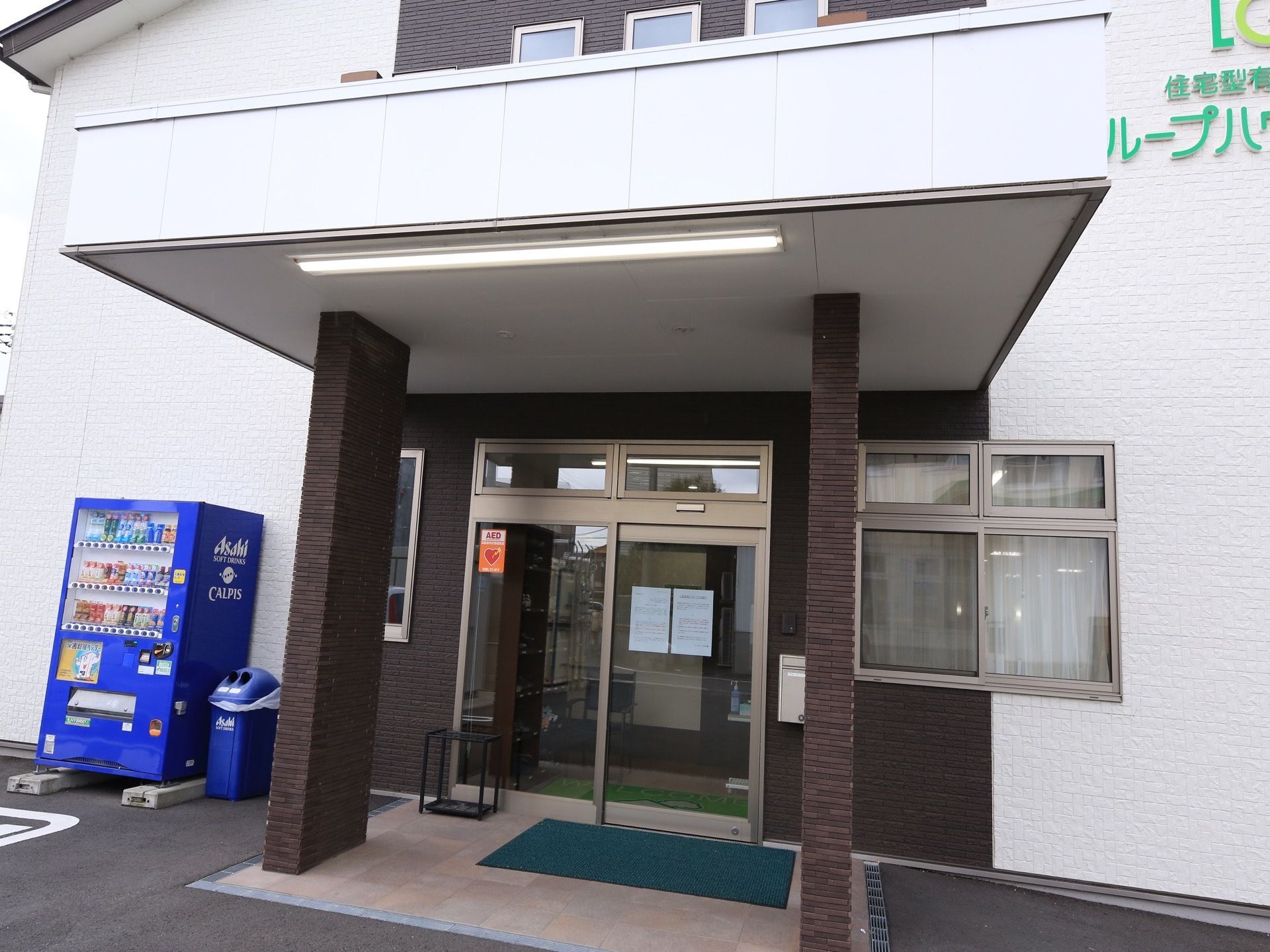 エントランスには、大きなひさしがついている。壁際に自販機が設置されており、ゴミ箱や傘立てが置かれている。
