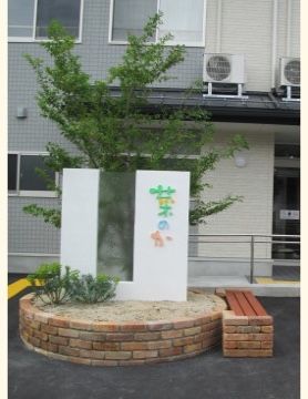 入り口近くにはシンボルツリーが植えられていて、施設名の看板が掲げられている。白い板にパステルカラーのロゴが柔らかい雰囲気。