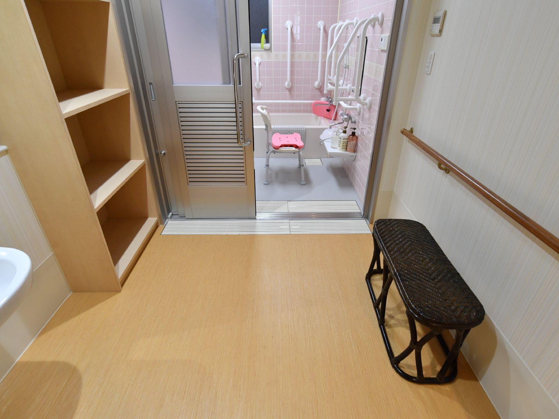 広さがあり、浴室との段差が無い。入口の近くには大きい棚が置かれている。壁には手摺があり、手摺の前に椅子がある。