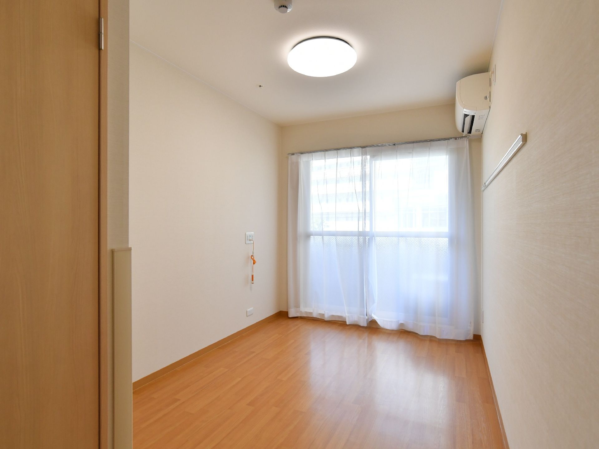 入口から撮影した、居室の写真。部屋の奥に窓があり、レースカーテンがついている。右上にエアコンが完備されており、左の壁にはナースコールがある。