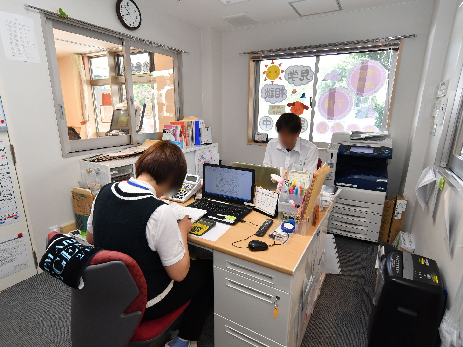 白色の天井と壁にグレイ色の床の中央にデスクが向かえ合わせに置いてある。パソコン、コピー機、収納棚などが置かれている。