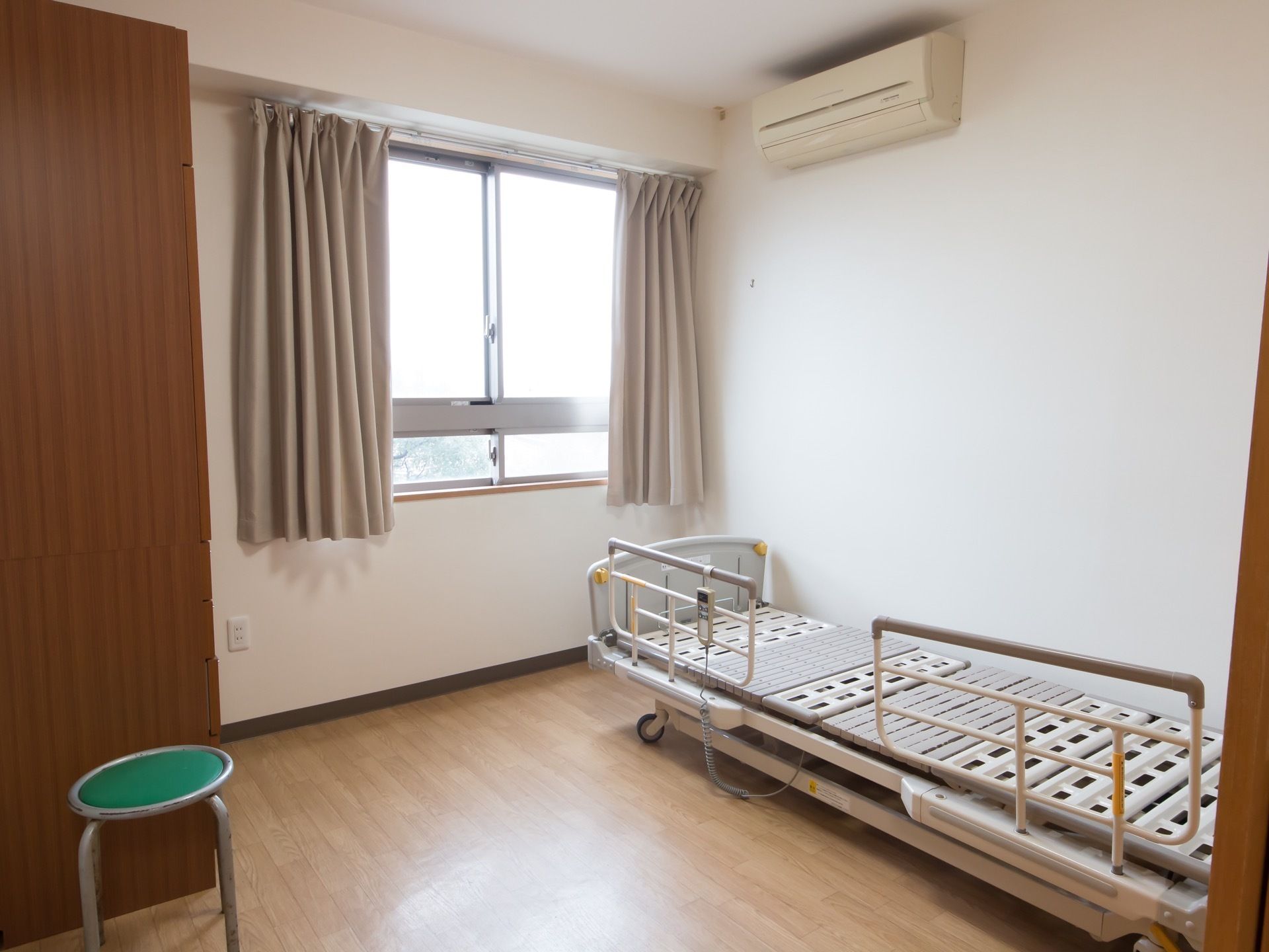 介護用電動ベッド、タンス、エアコンが備わった居室である。窓があり、ベージュのカーテンが掛けられている。