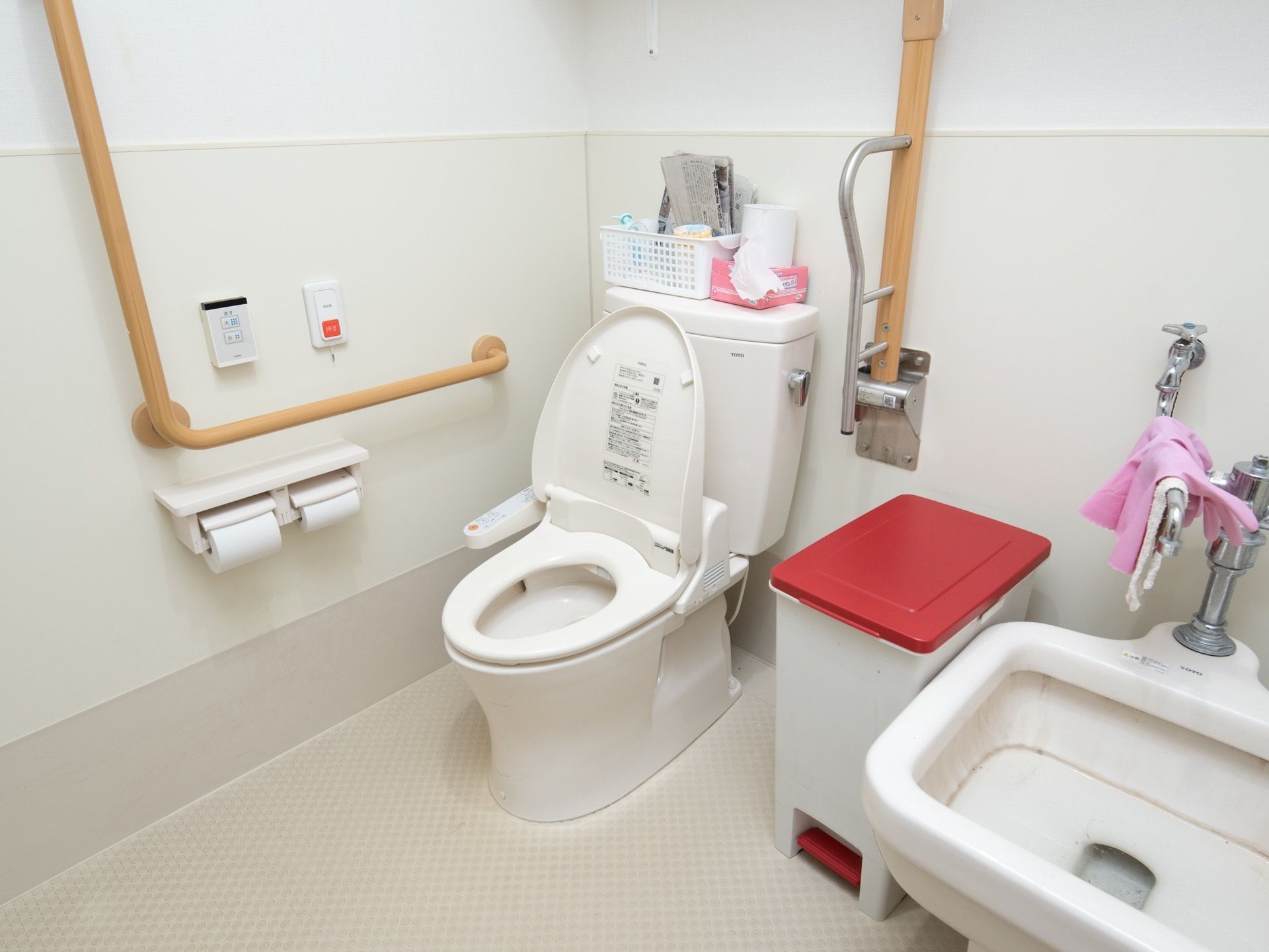 広々としたトイレ。壁には縦横方面に手すりが取りつけられており、便座には可動式のひじ掛けがついている。洗面台とゴミ箱がある。