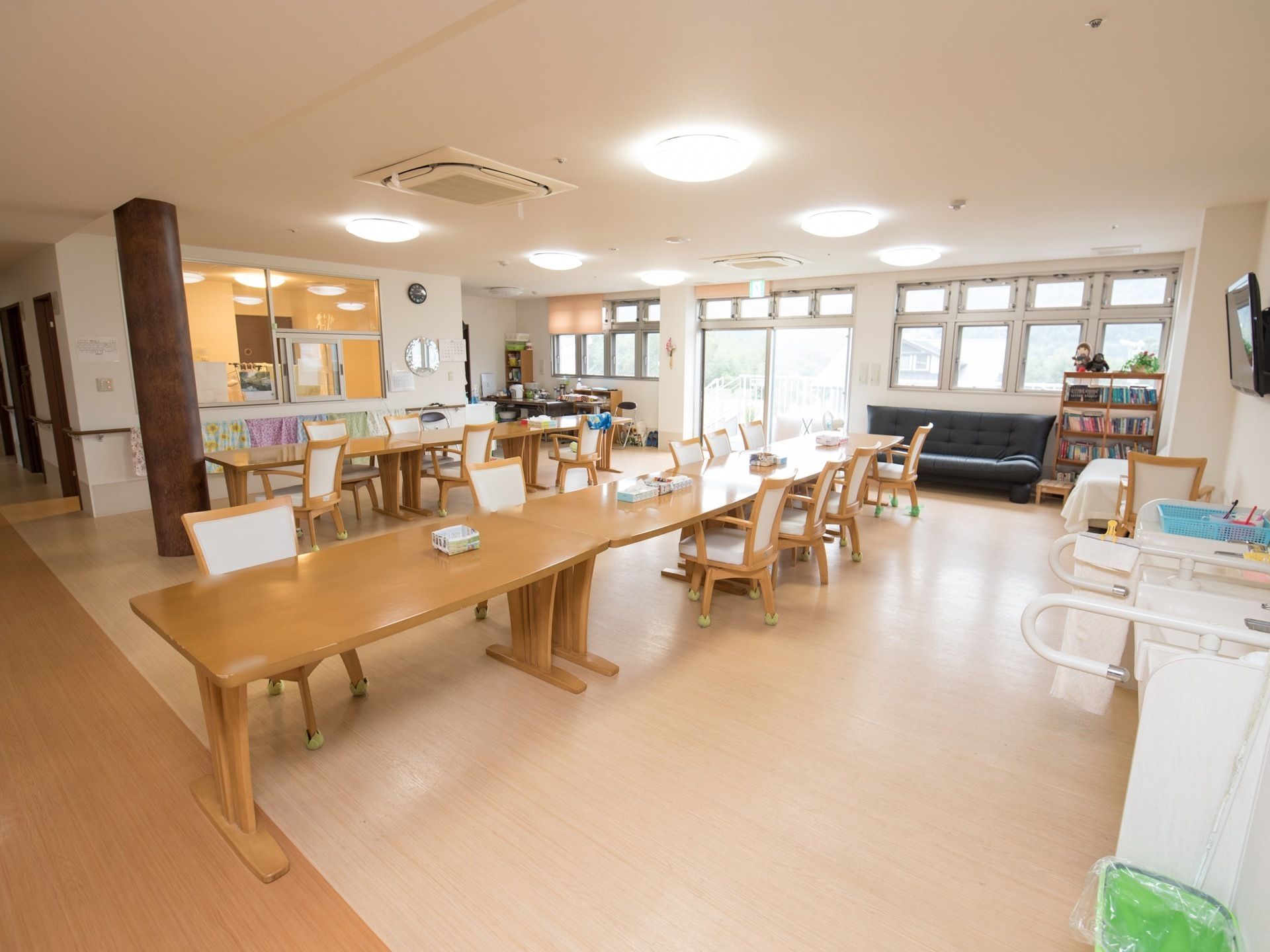大きなテーブルと椅子が並べられた食堂。窓が並び、外の景色が見える。壁際に洗面台と本棚、ソファが置かれている。