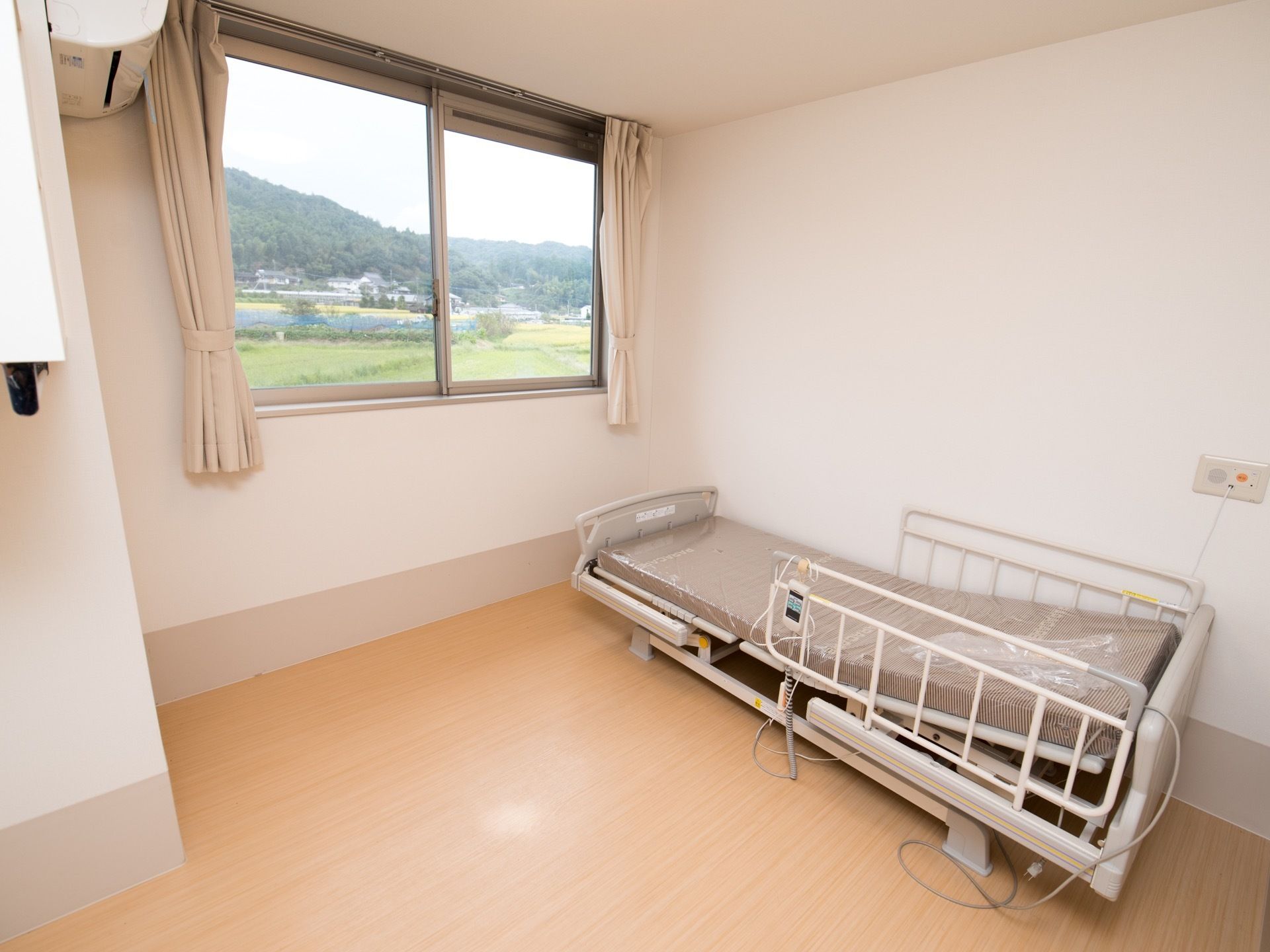 介護式ベッドが置かれた、落ち着いた色合いの入居者様の居室。大きな窓からは自然豊かな外の景色が見渡せる。