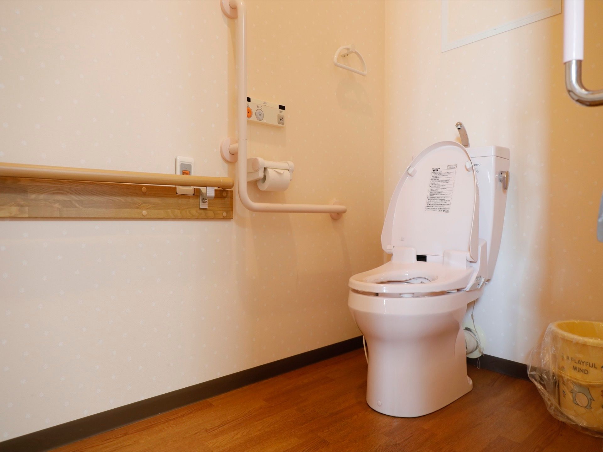 白い壁に木目調の床を取り入れたトイレである。便座の横の壁に、立ち座りの動作が楽に行えるようにＬ型の手すりを備えている。