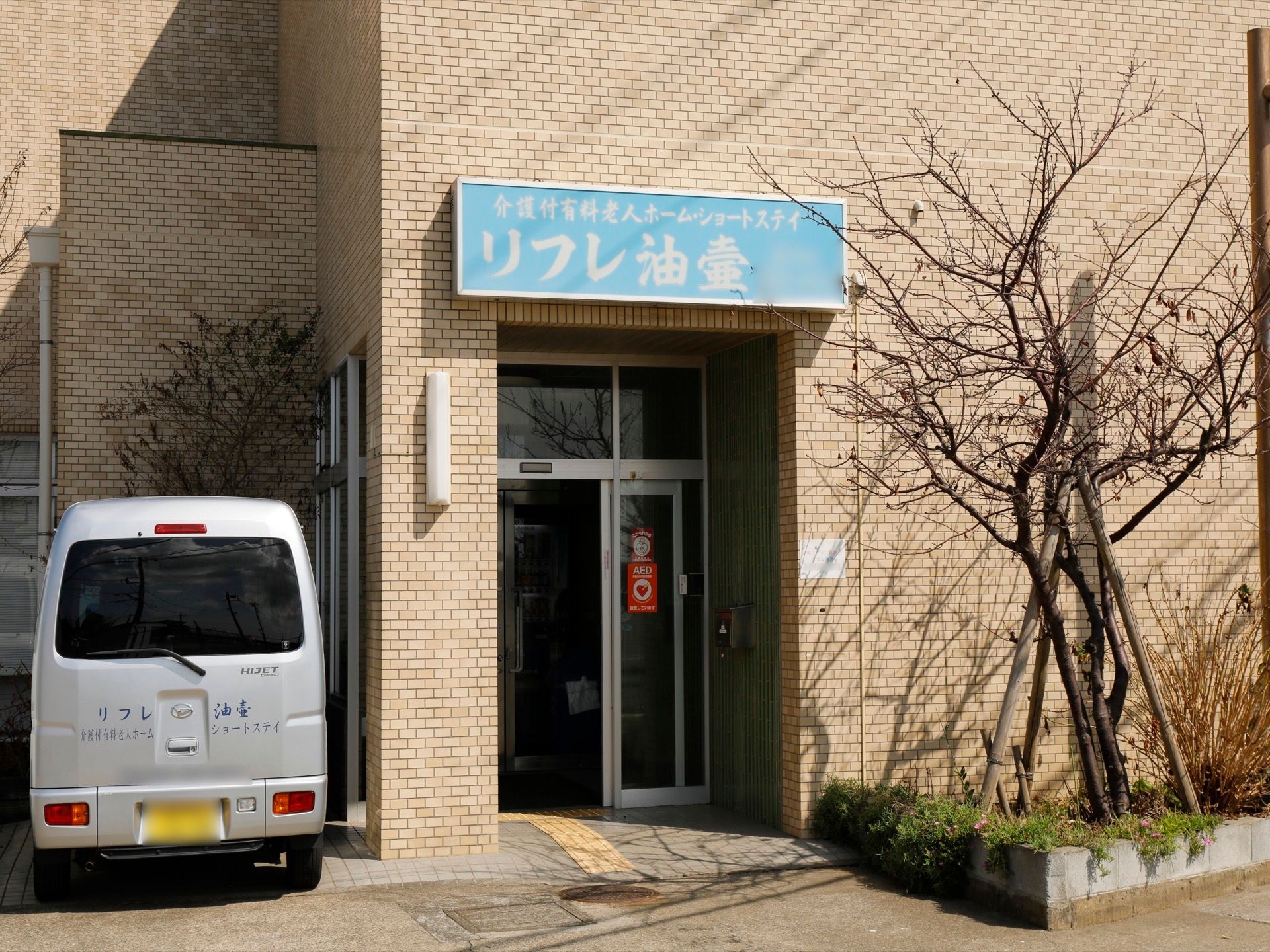 玄関ドア前の上部に、水色に白文字で、施設の看板を掲げている。その横側には樹木を植え、施設の専用車を停めている。