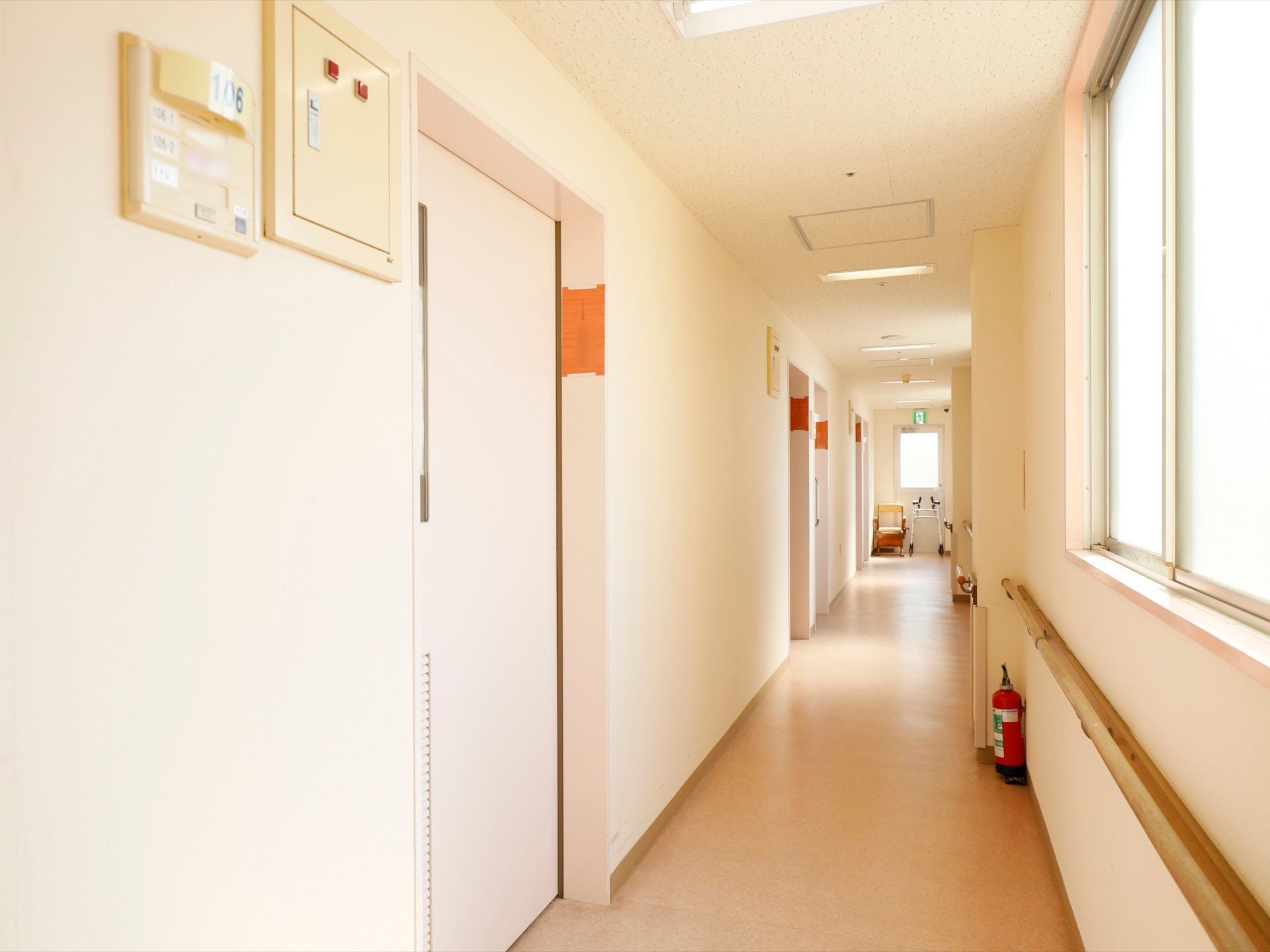 白を基調とした清潔感のある廊下。横側に窓があり、自然光が射し込む明るい空間である。壁際の床の上に消火器を備えている。
