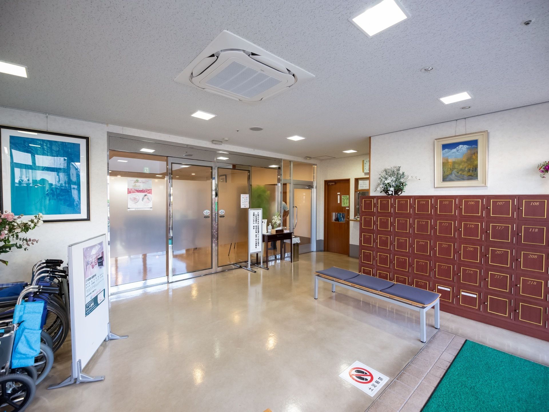 玄関内部に、居室ごとの靴箱が設置されている。土足厳禁の表示が床面に貼られている。靴を履き替えて、さらに自動ドアを通って施設内に入る。