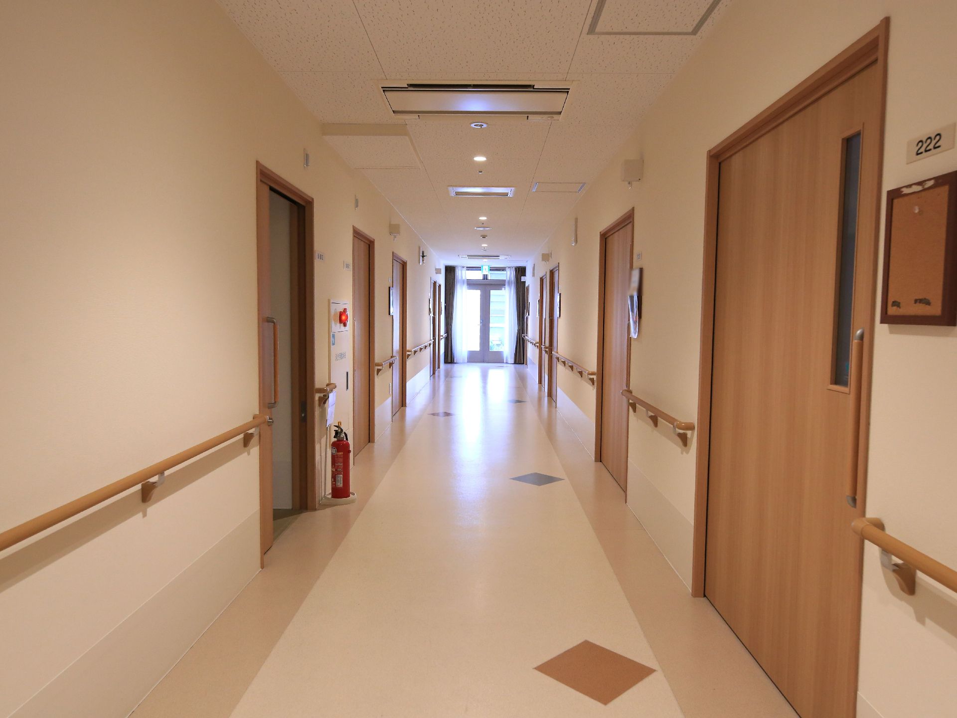 木目調のドアが並んでいる、ベージュの床の落ち着いた雰囲気の廊下。壁には手すりが設置されており、突き当たりには大きな窓がある。
