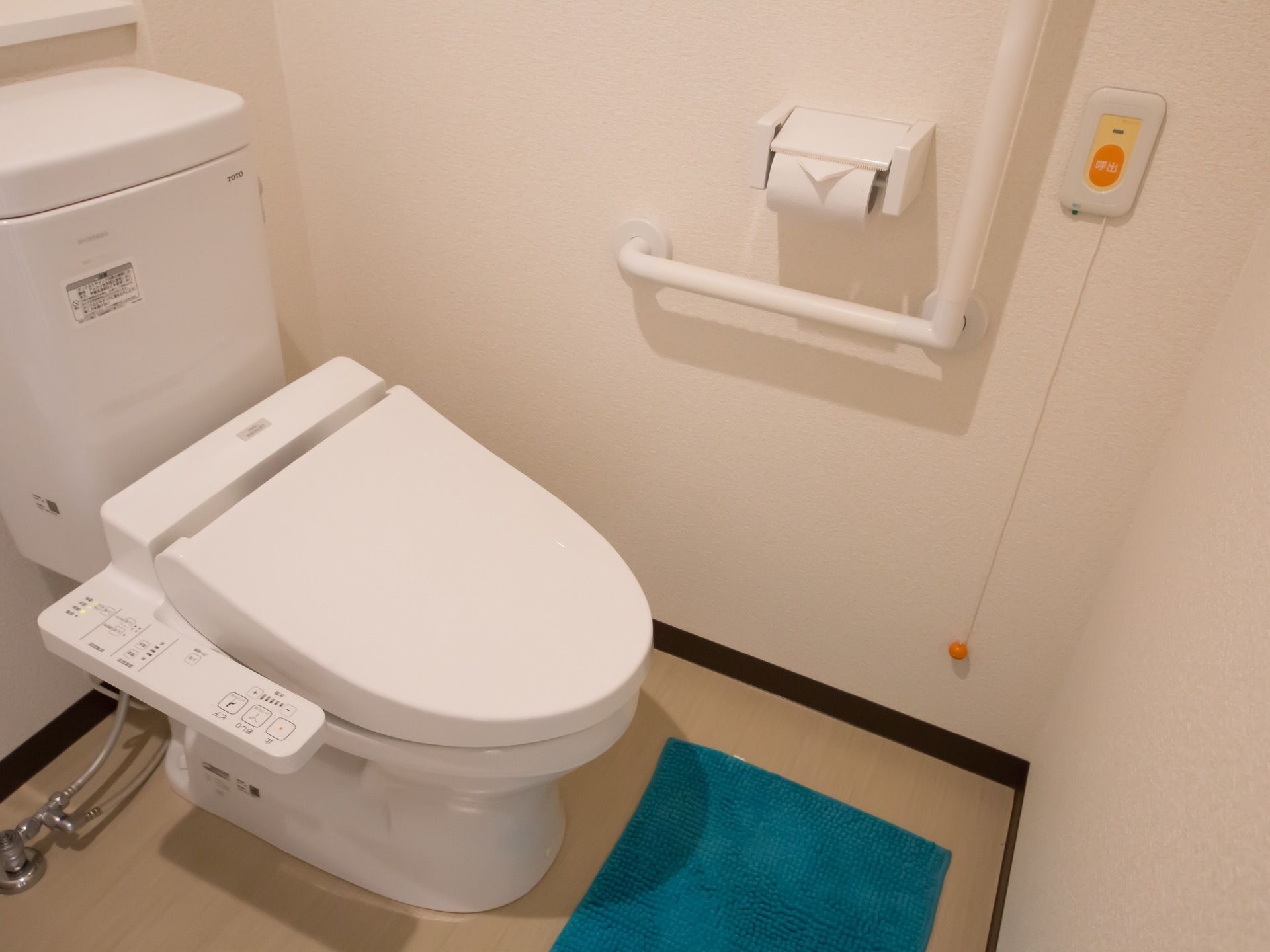 壁にＬ字型の手すりや紐の付いたナースコールが取り付けられている。床に青いトイレマットが敷かれている。