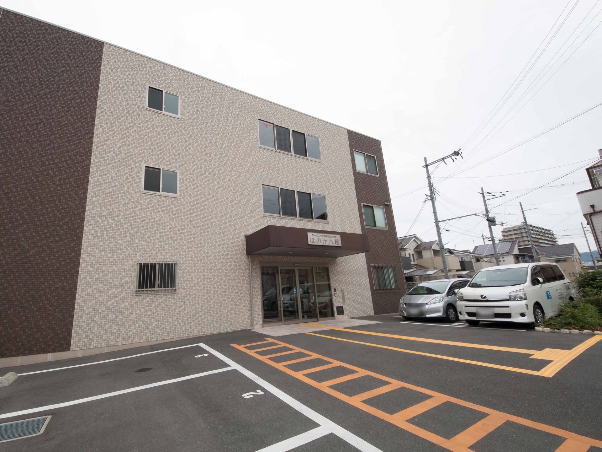 ３階建てのブラウンを基調とした四角いグループホームである。屋根が付いた玄関の前には点字ブロックが設置されている。