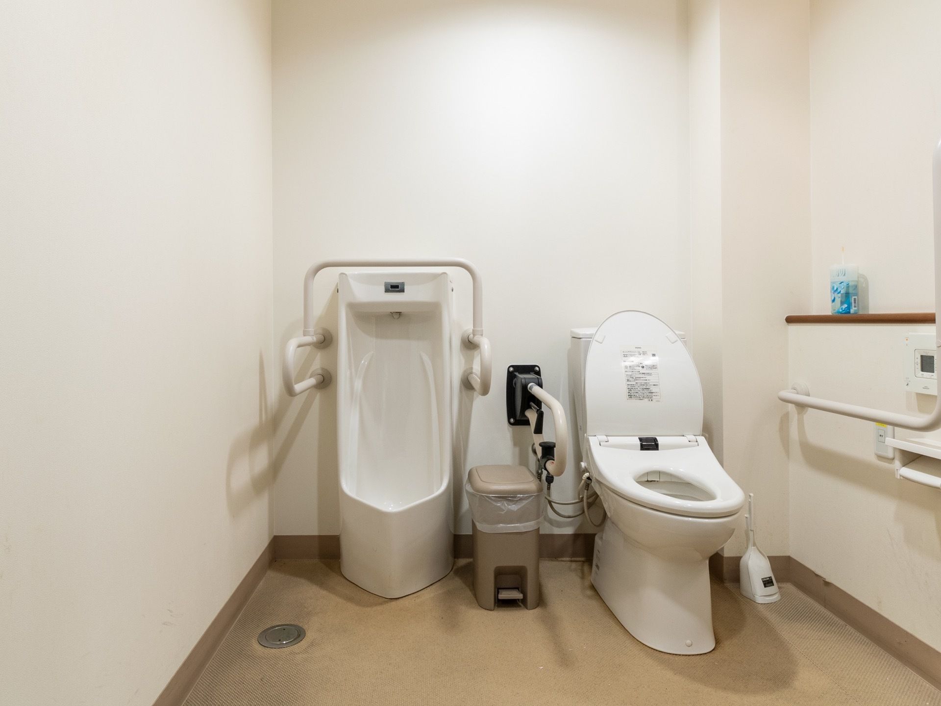 男子用小便器と大便器が一緒の部屋に設置されている共用のトイレ。両方ともにパイプ製の手すりが取り付けられている。