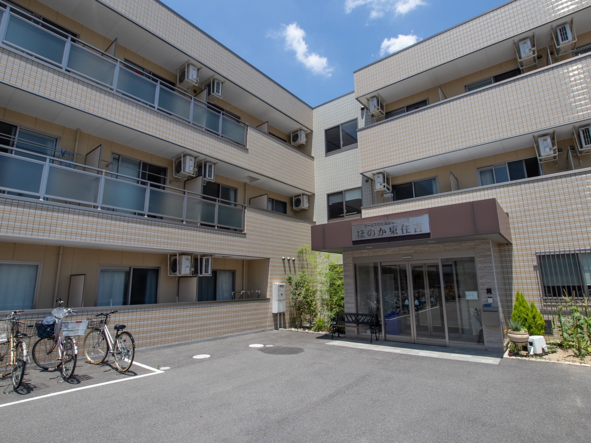 白いタイル調の３階建てのエル型構造の外観である。玄関前は広々としたスペースを設け、横側に駐輪場を備えている。横側に緑の花壇がある。