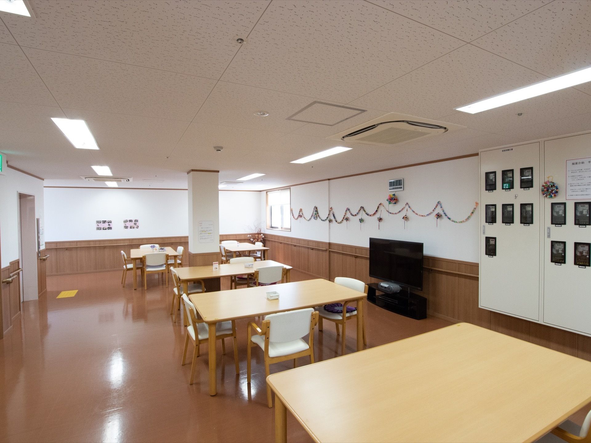 ダイニングテーブルと白い座面に背もたれが付いた椅子が設置されている。壁際にはテレビが置かれ、壁には折り紙で作られた飾りが飾ってある。