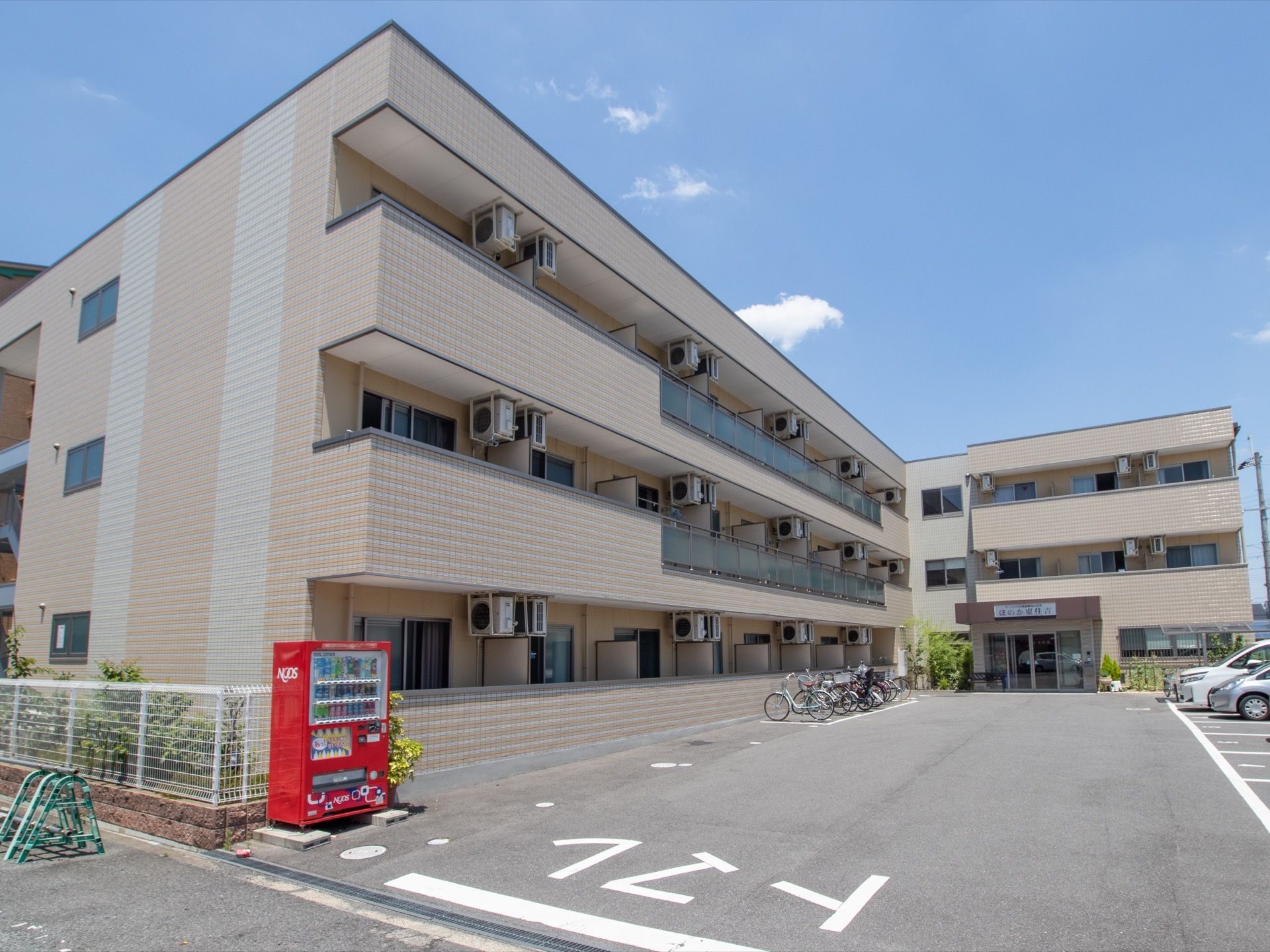 白い壁で3階建てのマンション型施設である。入口に自動販売機が置かれている。駐車場、駐輪場も完備されている。