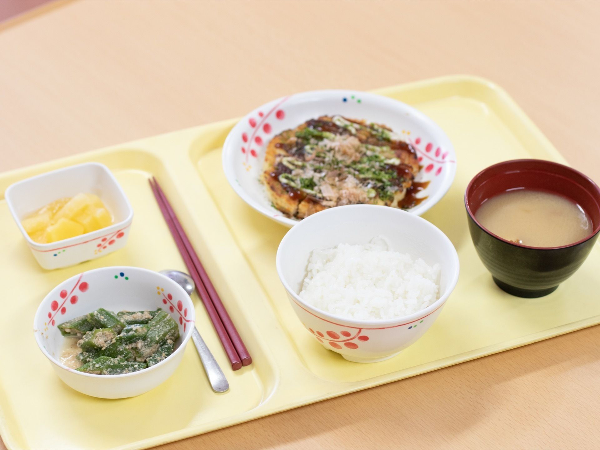 プラスチックトレーの上にお箸、スプーン、白米、みそ汁、お好み焼き、和え物、フルーツが用意されている。