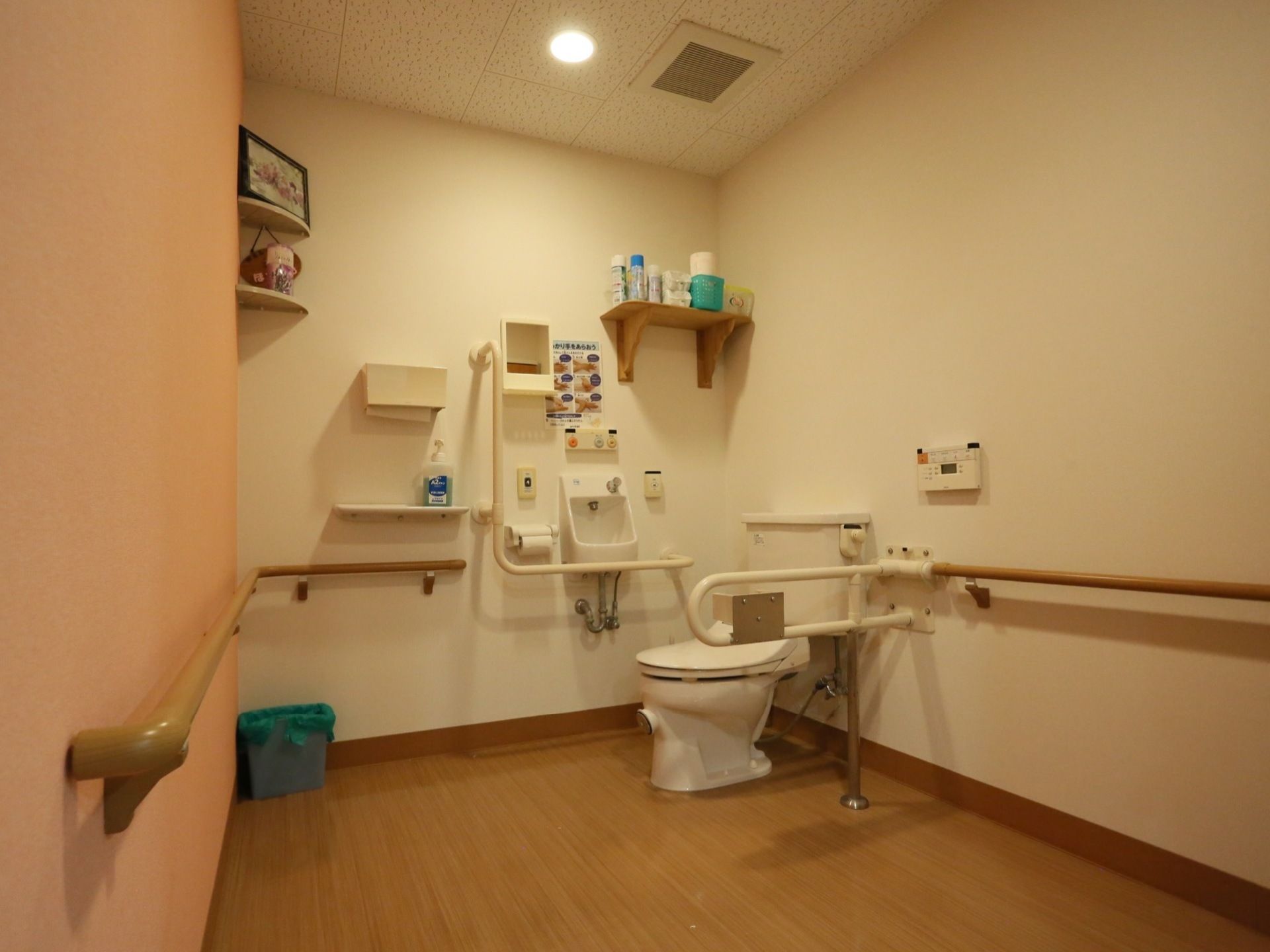 広いトイレ内には、両側に手すりのついた洋式便器があり、脇に小さな手洗い場が設置されている。壁にはペーパータオルや収納棚が備えつけられている。