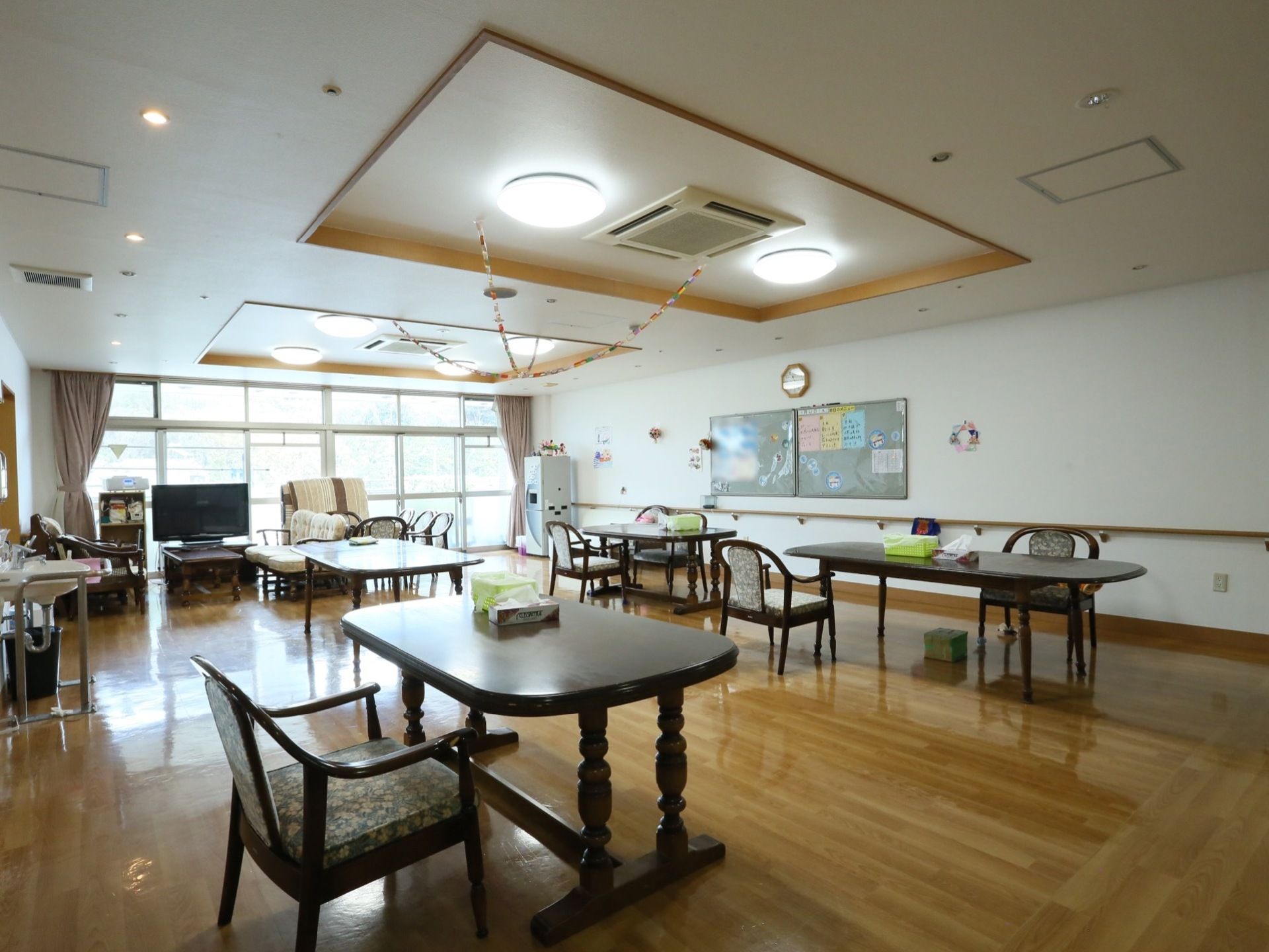 フローリングの部屋にダークブラウンのダイニングテーブルが並んでいる。奥にはテレビやソファ、手洗い場が設置されている。