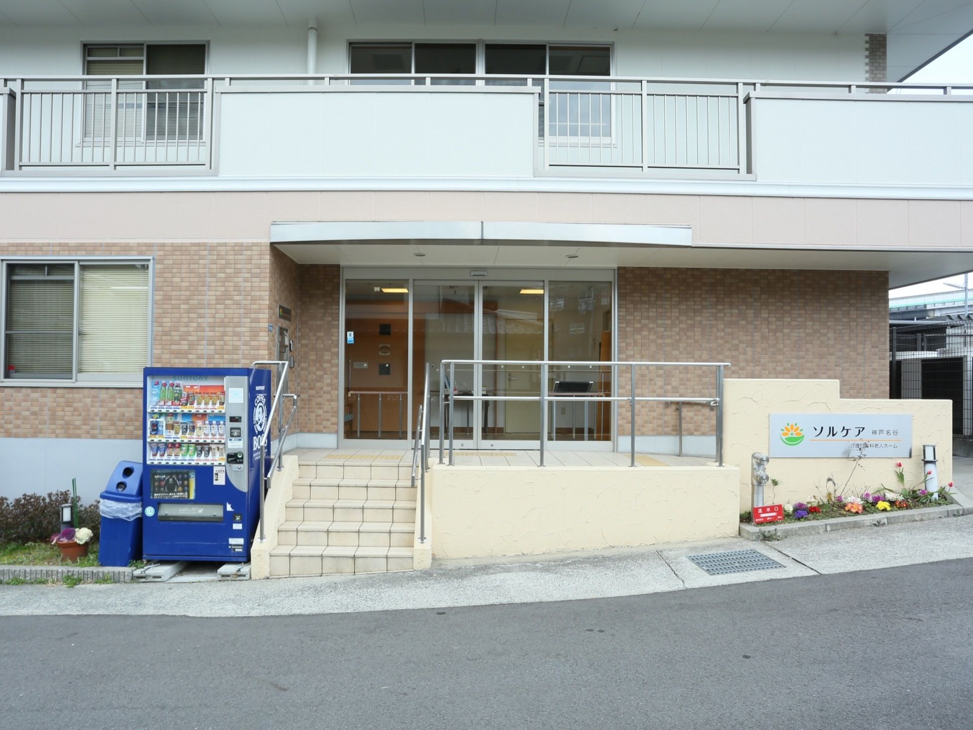 玄関前には階段とスロープ両方を備えている。施設名のプレートの下には小さな植え込みがあり、花が咲いている。