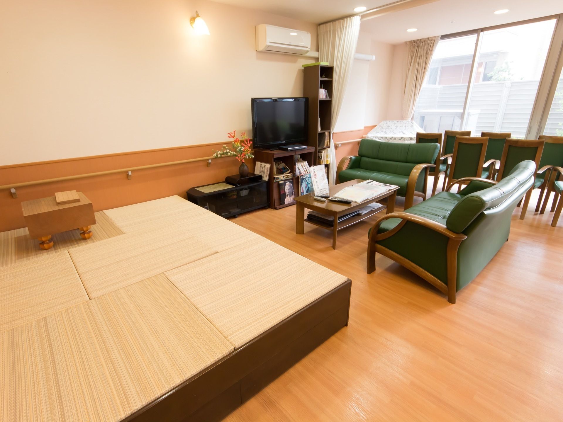 テレビの前に長テーブルが置かれており、囲むように緑のソファーが設置されている。テレビの左側に畳が敷かれたスペースがある。