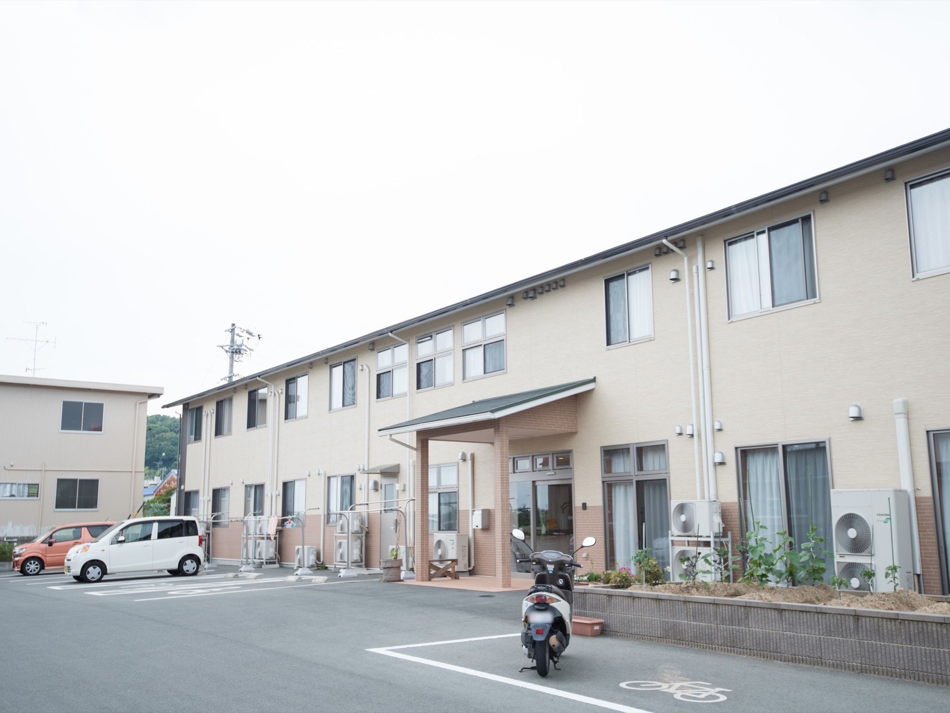 淡いブラウンが基調の２階建ての建物である。周辺には物干し台や植え込み、駐車スペースが完備されている。