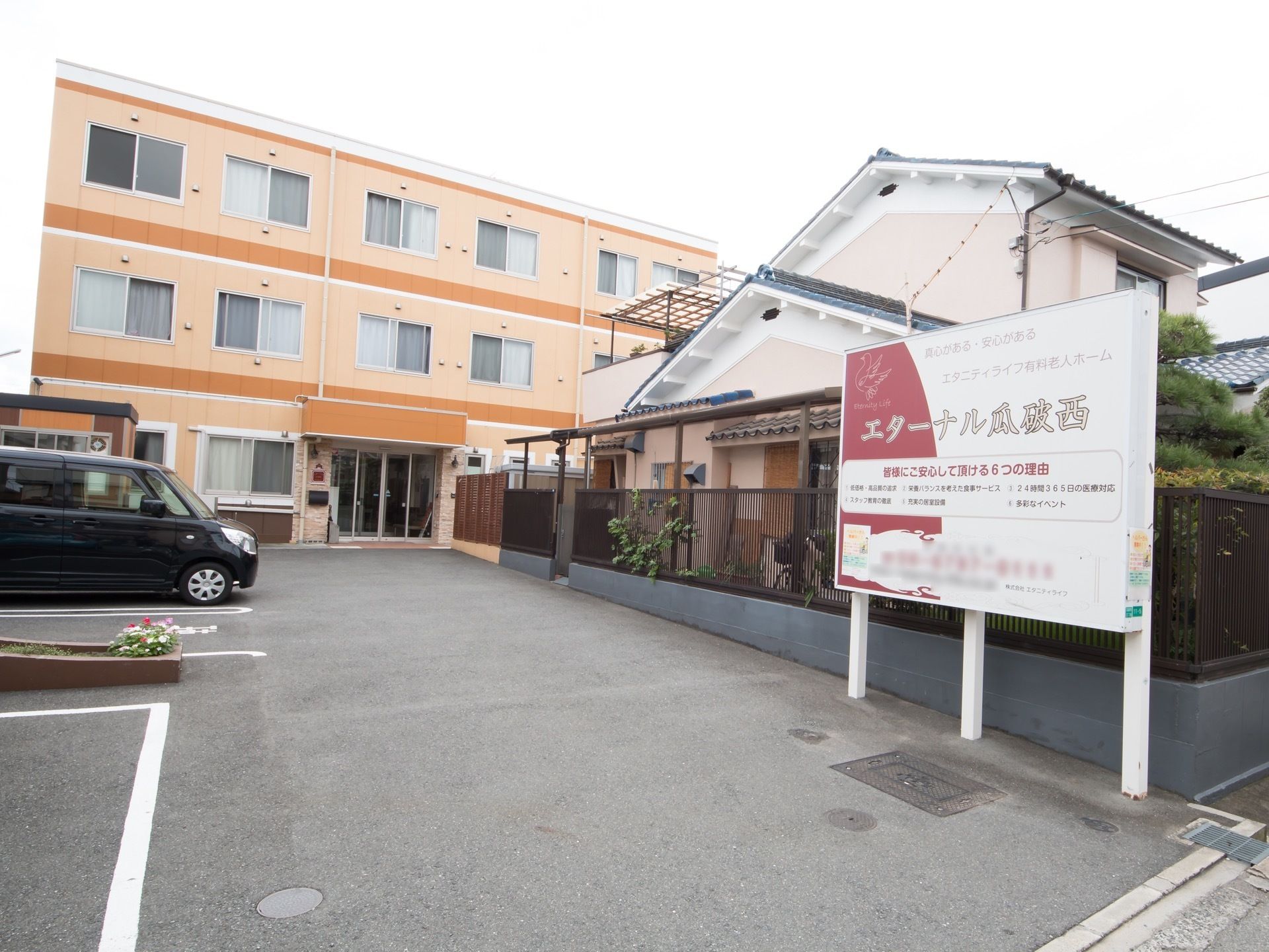 3階建ての建物で、施設前は駐車スペースになっている。道路側に大きな看板が立っており、施設名などが書かれている。