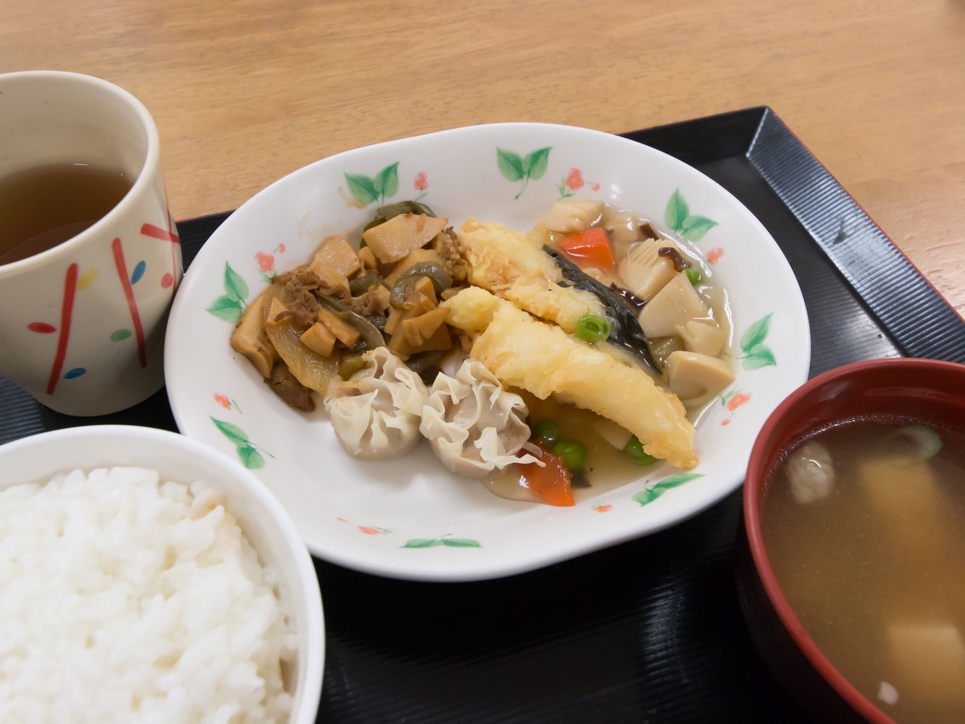 黒いトレイの上には、ご飯や味噌汁、３種類のおかずが盛られた一皿が並べられている。お茶の入った湯呑も置かれている。