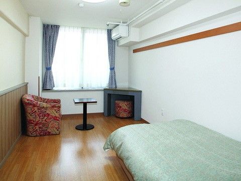 奥行きある広々した居室である。手前にはグリーン系の布団カバーがつけられたベッド、奥に１人掛けソファとテーブルがある。