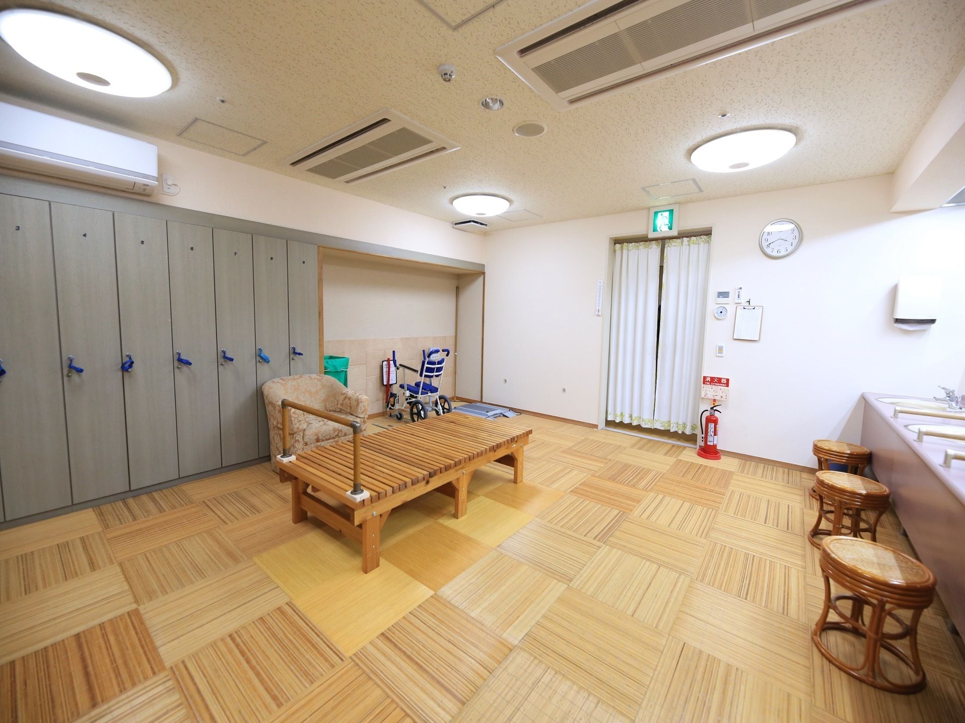 落ち着いた内装で、明るく広々とした脱衣所である。洗面台と鍵付きの個別ロッカーが完備されている。中央に手すり付きのベンチが置かれている。