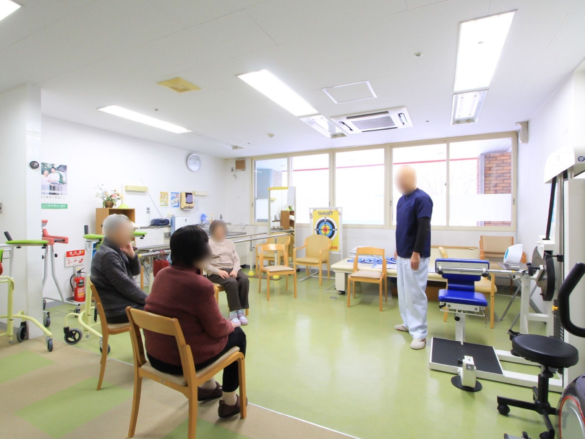 歩行訓練用の手すりや椅子、バイクなどが設置されたフロアがある。入居者様が数名椅子に座って、スタッフと話をしている。