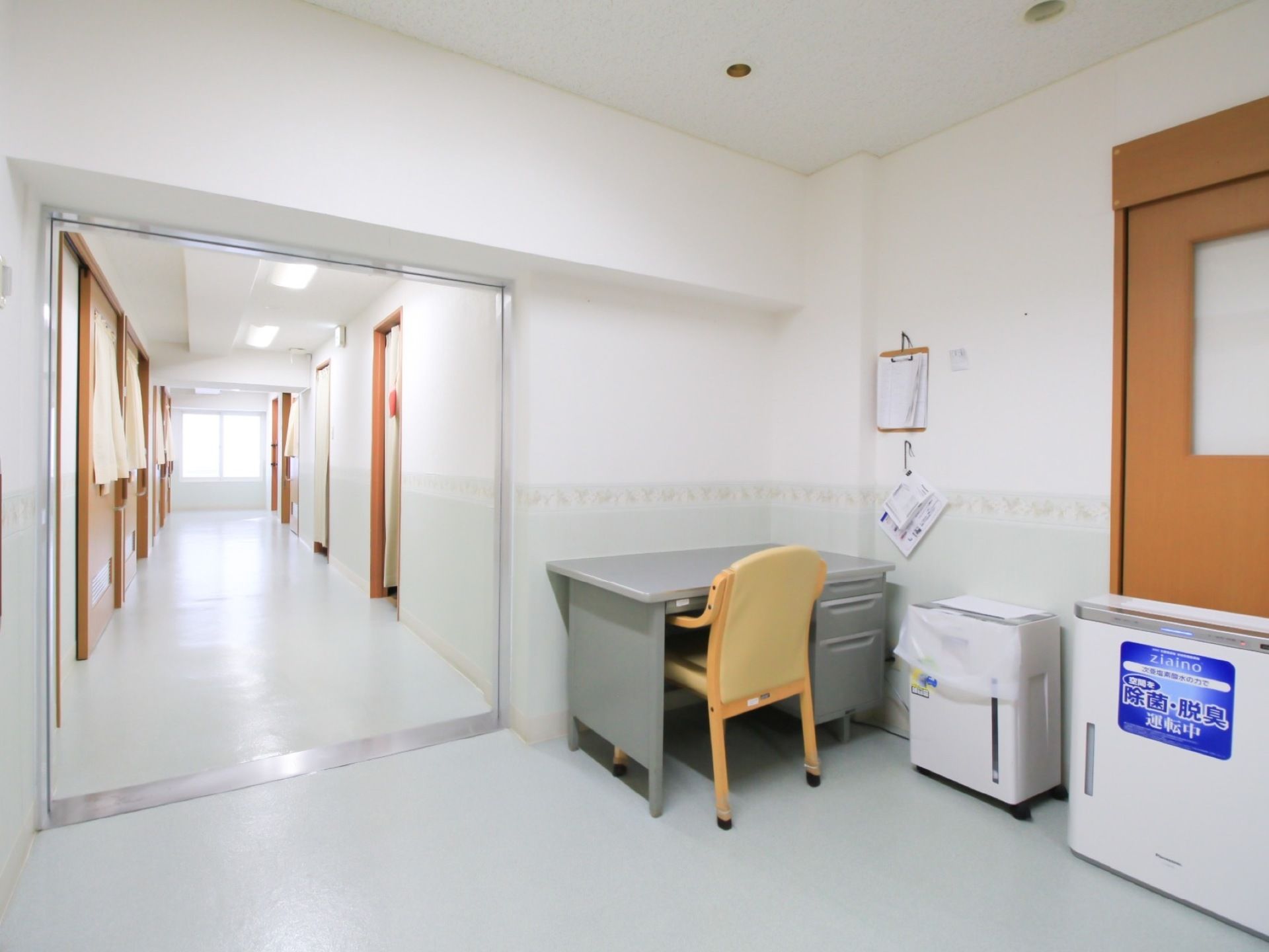 白いビニール床の廊下が続いている。一角に執務デスクと椅子があり、その横には空気清浄機が設置されている。