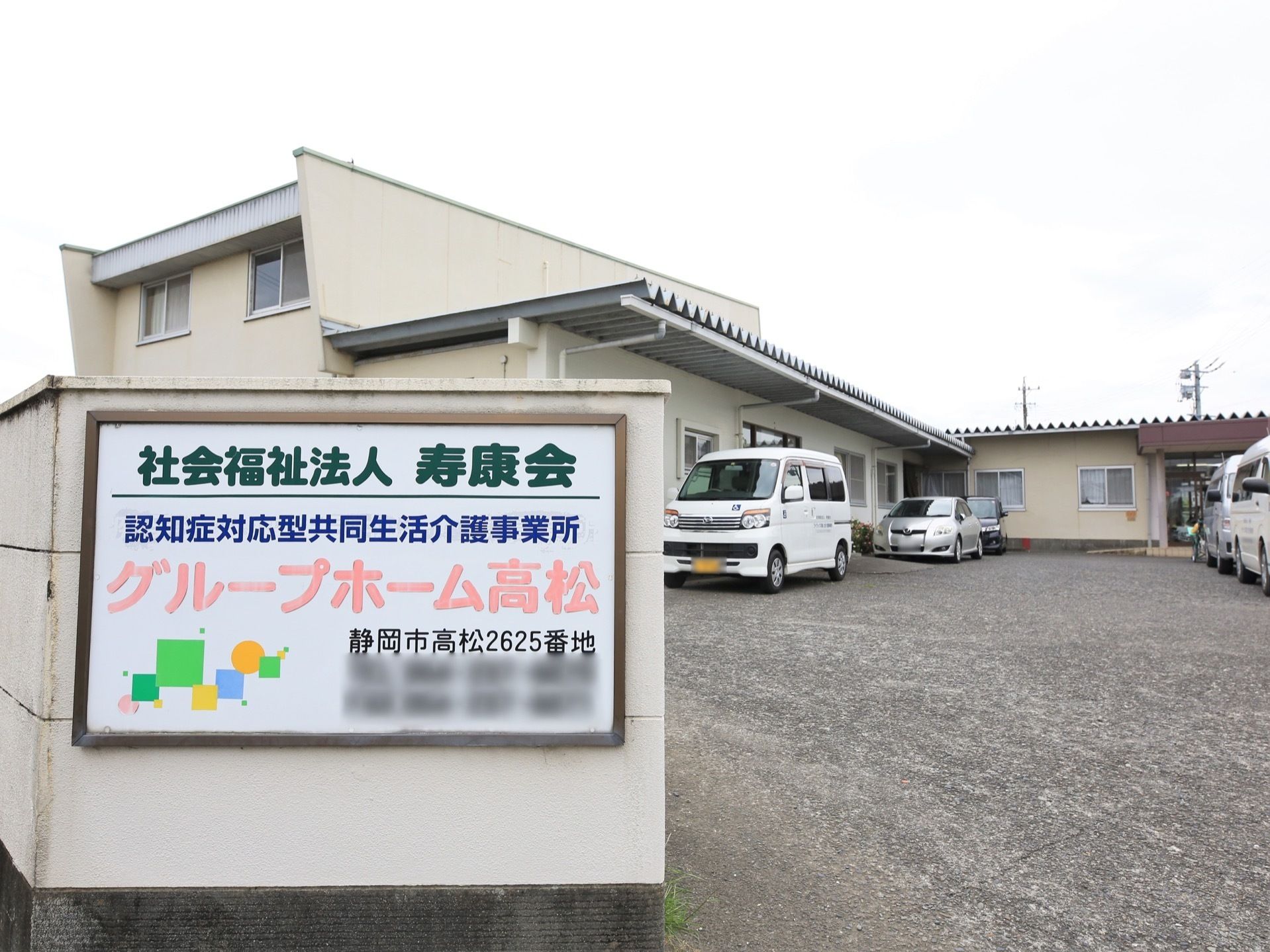 塀に四角い看板が設置されており、法人名や施設名称が明記されている。塀の内側には、Ｌ字型の建物と駐車スペースがある。