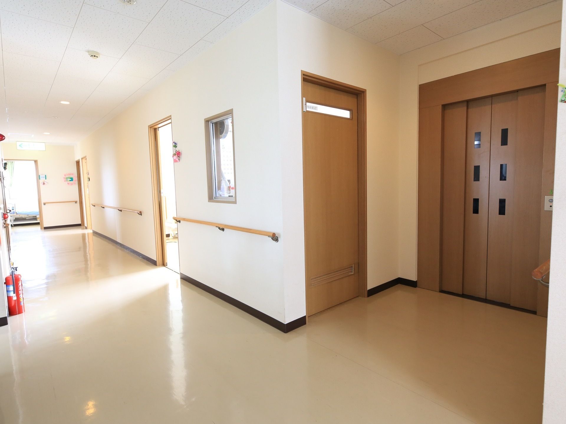 木製の手すりが連続する通路の一角に、エレベーターが見える。扉やその周囲は、落ち着いた色味の木目調である。