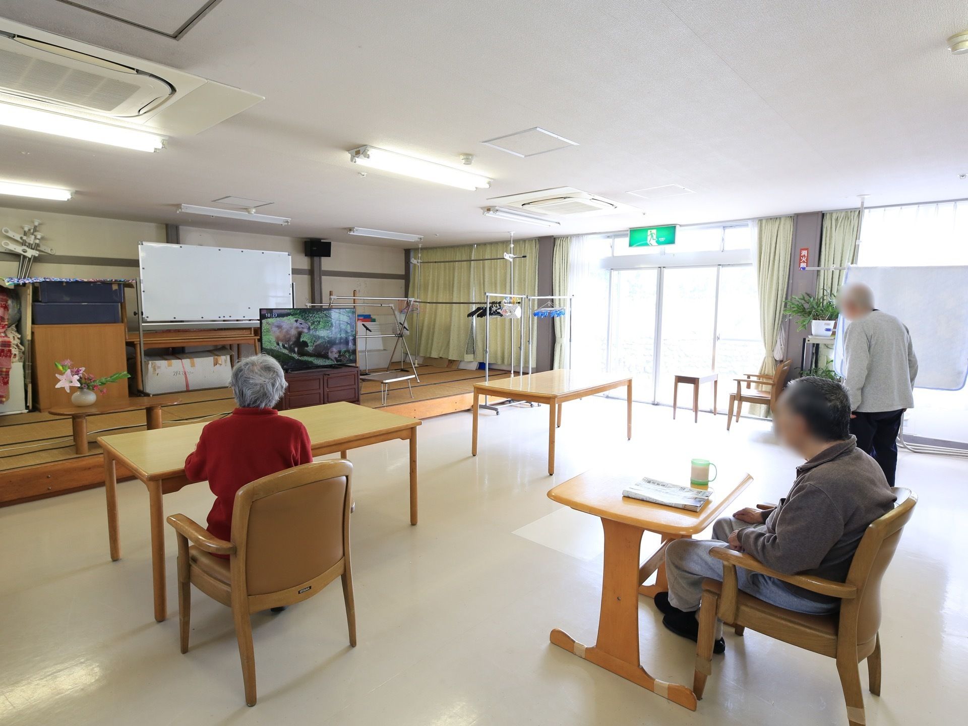 右奥には物干し台が数種類用意されており、窓側にシーツが干されている。テレビを観ている方々の姿が見える。