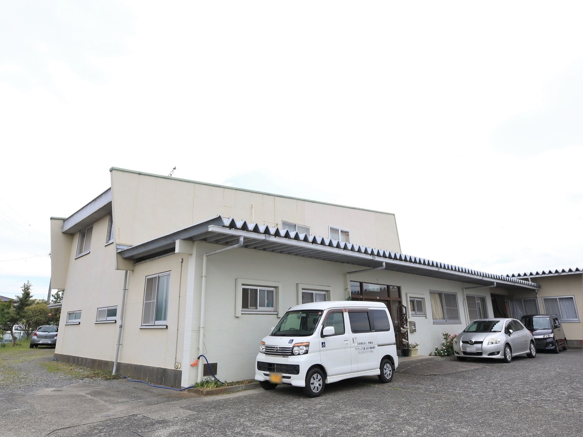 Ｌ字型の建物の周囲には舗装されたスペースが広がっており、複数の自動車が停められている。木々の緑や花壇の花が見える。