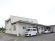 Ｌ字型の建物の周囲には舗装されたスペースが広がっており、複数の自動車が停められている。木々の緑や花壇の花が見える。