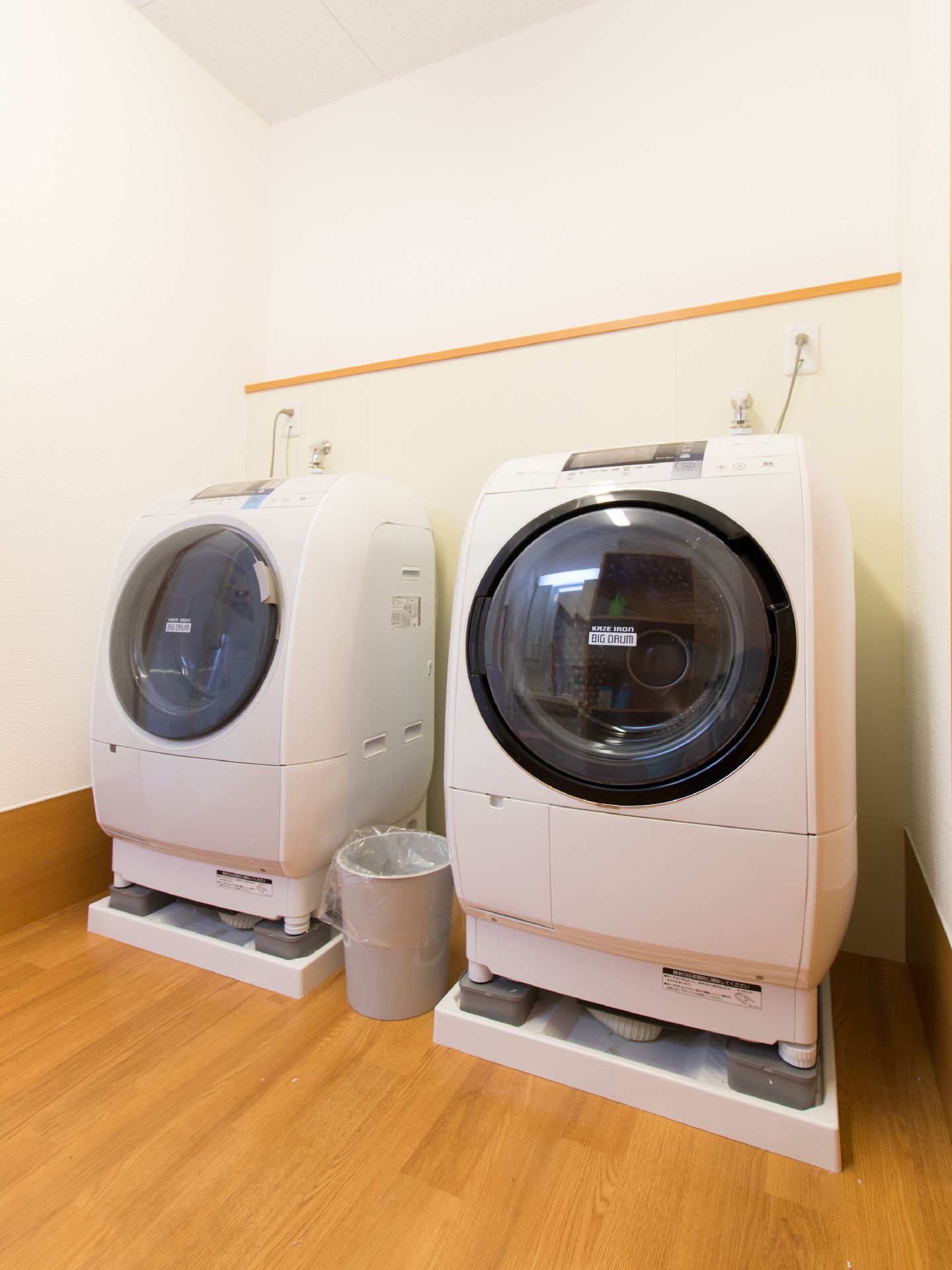 大きな洗濯機が2機並んでいる。いずれもドラム式である。その中央にはごみ箱がある。床は木目調で壁は白い。