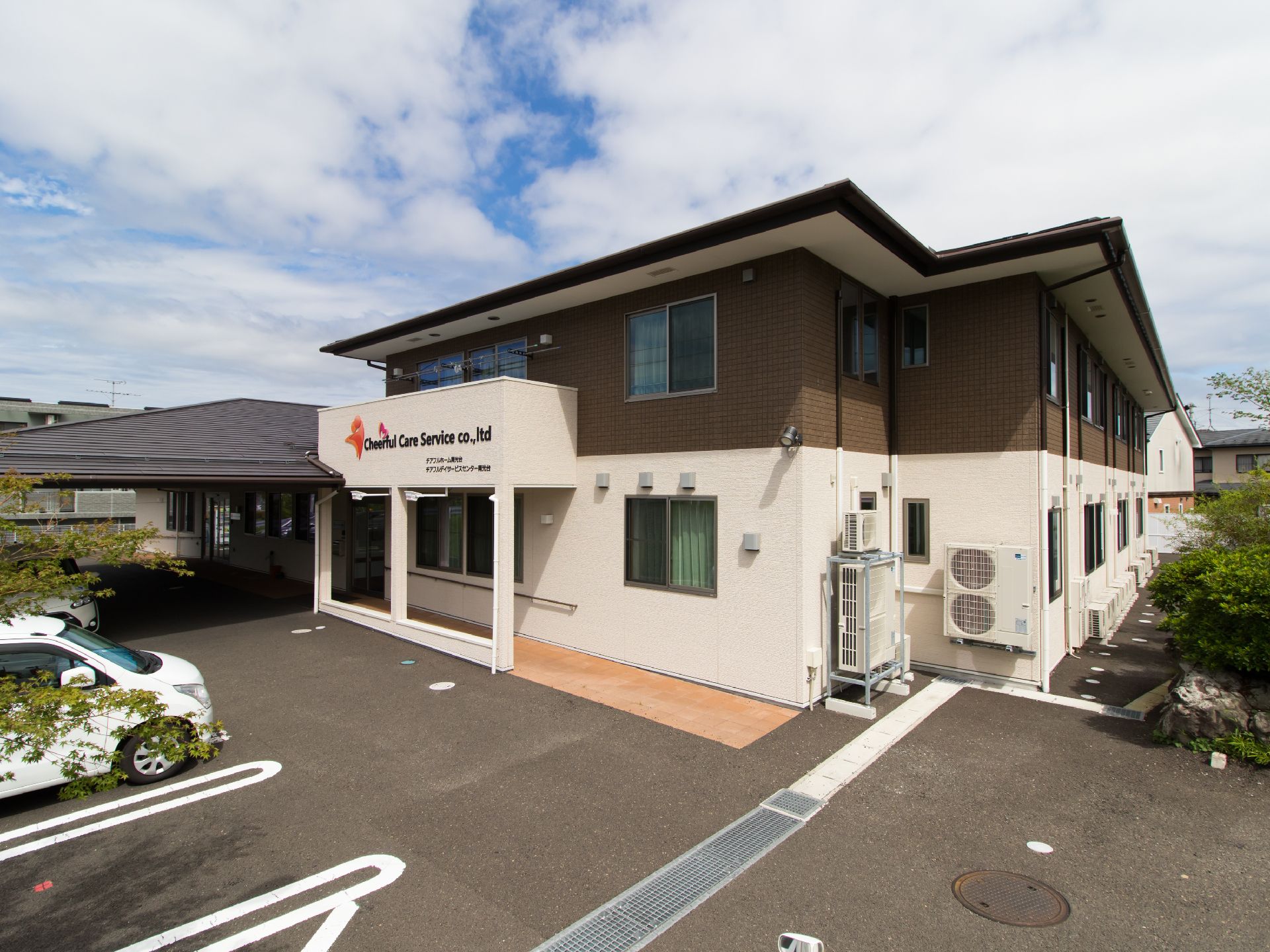 玄関には施設名が見える。建物は茶色と白のツートンカラーで構成されている。駐車場には自動車が駐車している。