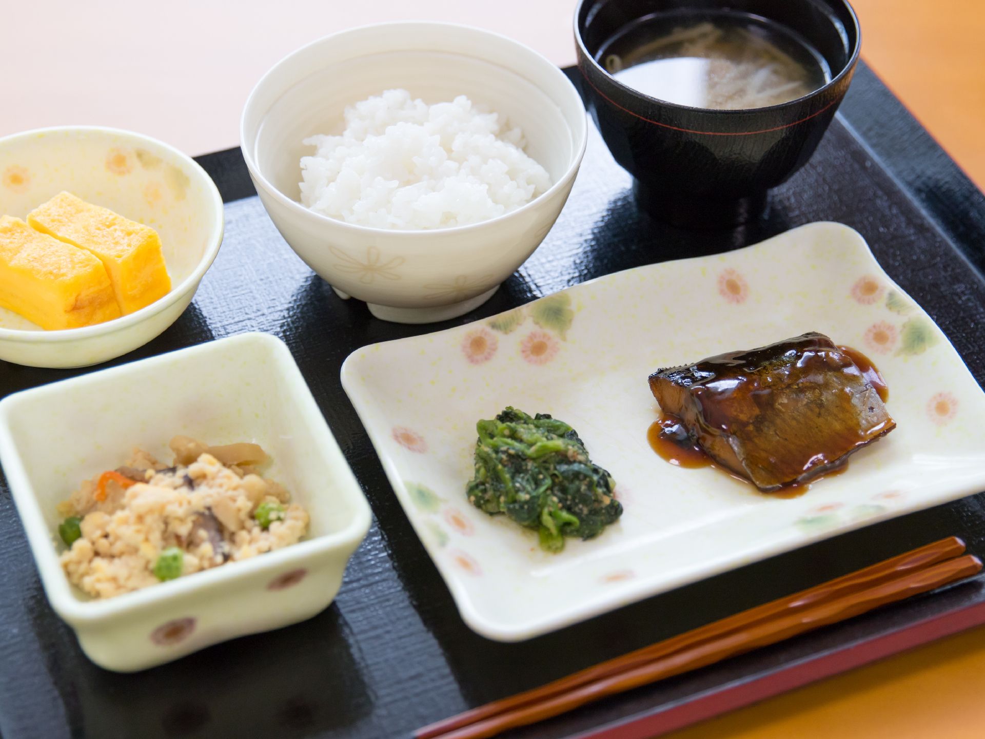 和食が黒い盆に載っている。米飯と汁物に、卵焼きと魚料理、おからと青菜をそれぞれに利用した副菜がある。木の箸が添えられいる。