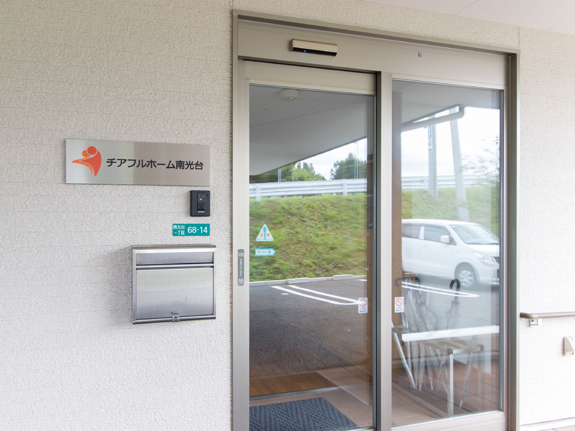 施設名が表示された小さめのプレートがある。郵便受けがあり、その上にインターホンが設置されている。ガラス張りの自動ドアに自動車が映っている。