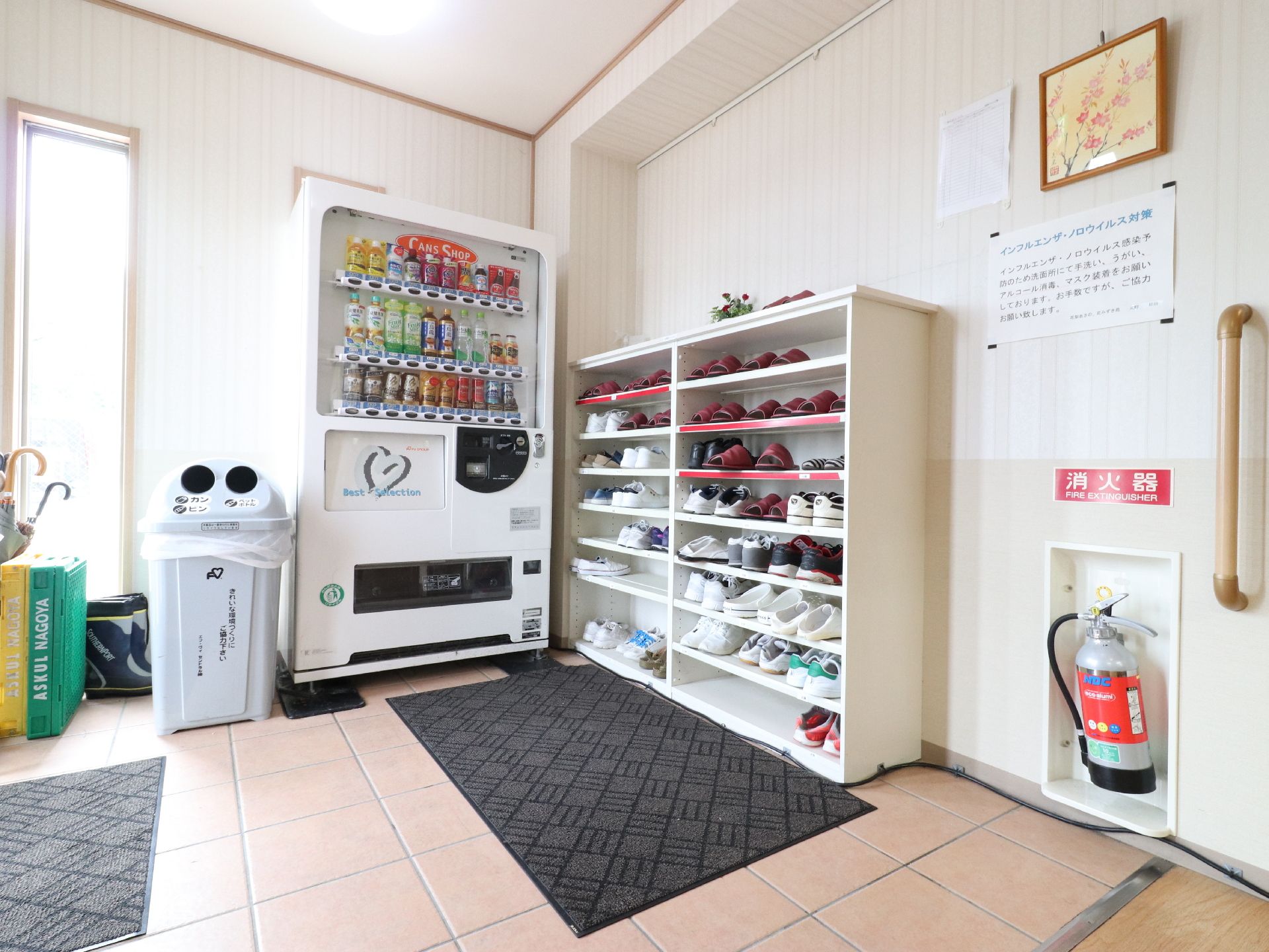 床がタイルの玄関には、マットが敷いてある。自動販売機があり、飲み物が売っている。上がり框に靴箱があり、靴やスリッパが置いてある。