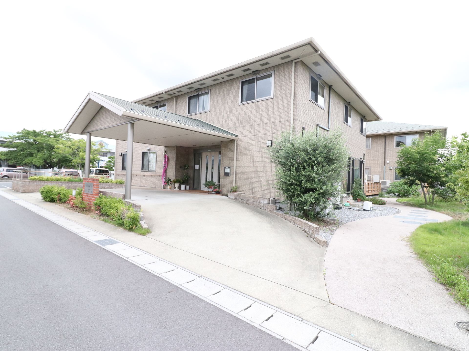 歩道そばは低木が植えてある。施設横に駐車場がある。反対側の横には、遊歩道が設けてある庭がある。玄関前にプランターがある。