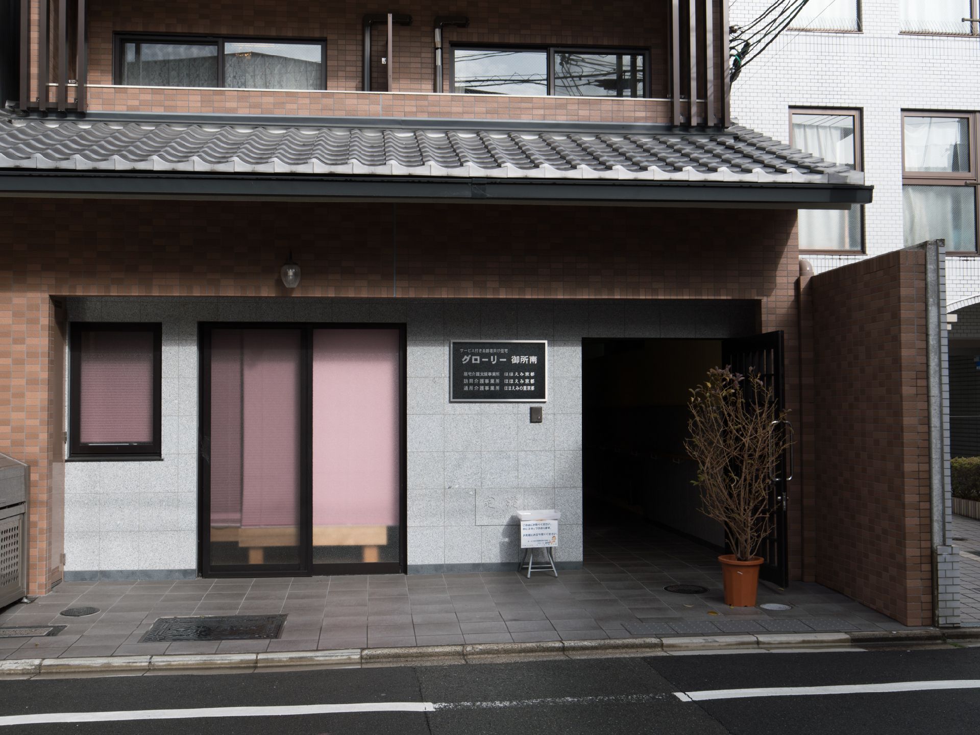 通りに面した建物の1階はガラスサッシと、エントランスに通じる入り口があり、正面に建物名の表札が付いている。植木鉢が置かれている。
