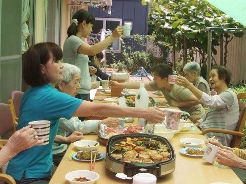 庭先に長机を出しホットプレートを囲みながら楽し気に乾杯の動作をするスタッフと入居者たち