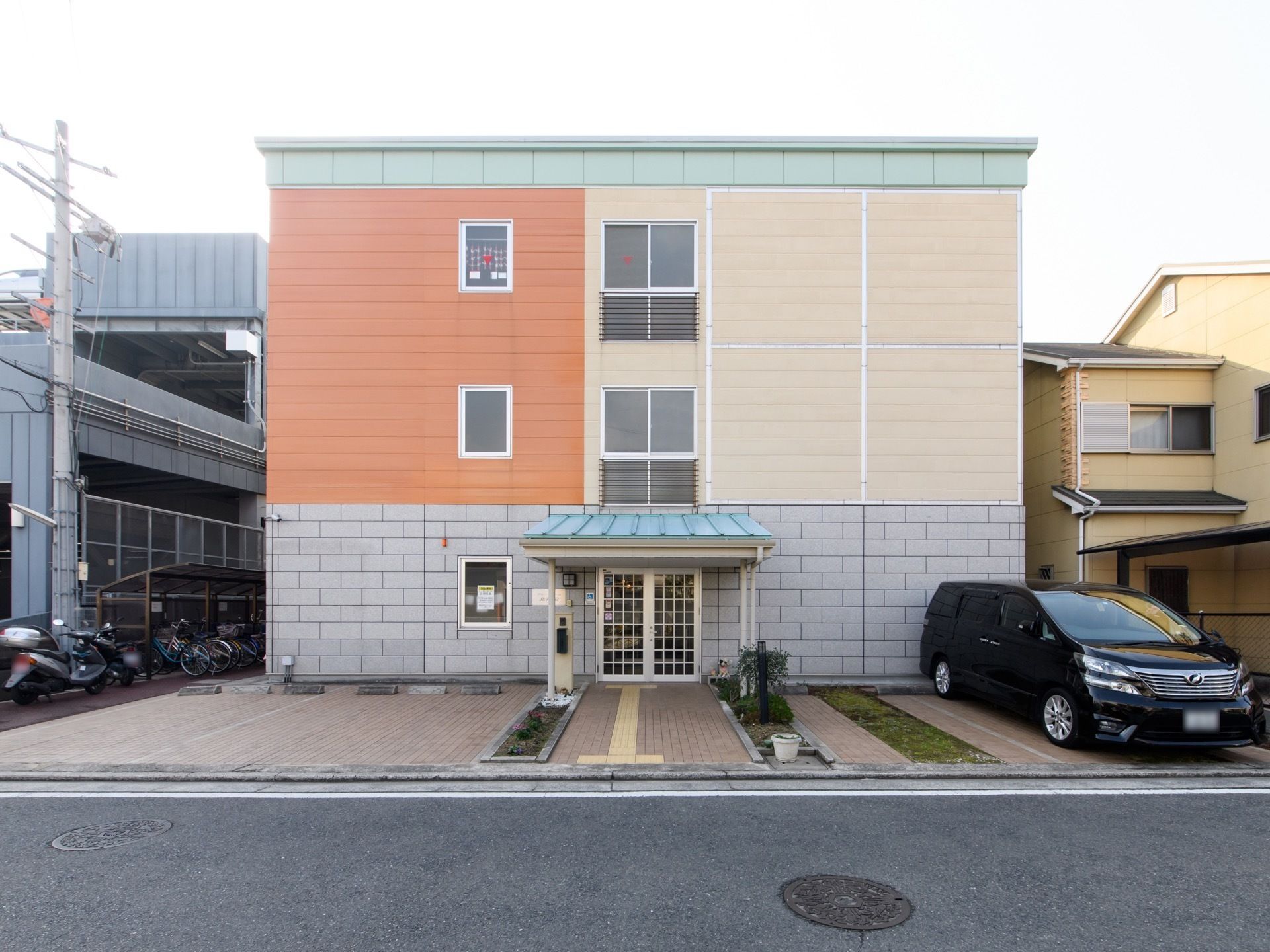 淡い色の配色の３階建ての建物で、施設前に駐車場が完備されている。駅近くに立地されている。入口前にひさしがある。