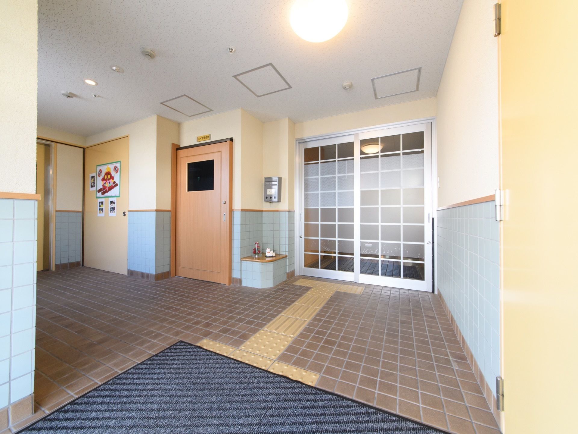 ガラス製の入口ドアである。郵便受けが掛けられている。玄関前の広々としたホールは屋根が取り付けられている。