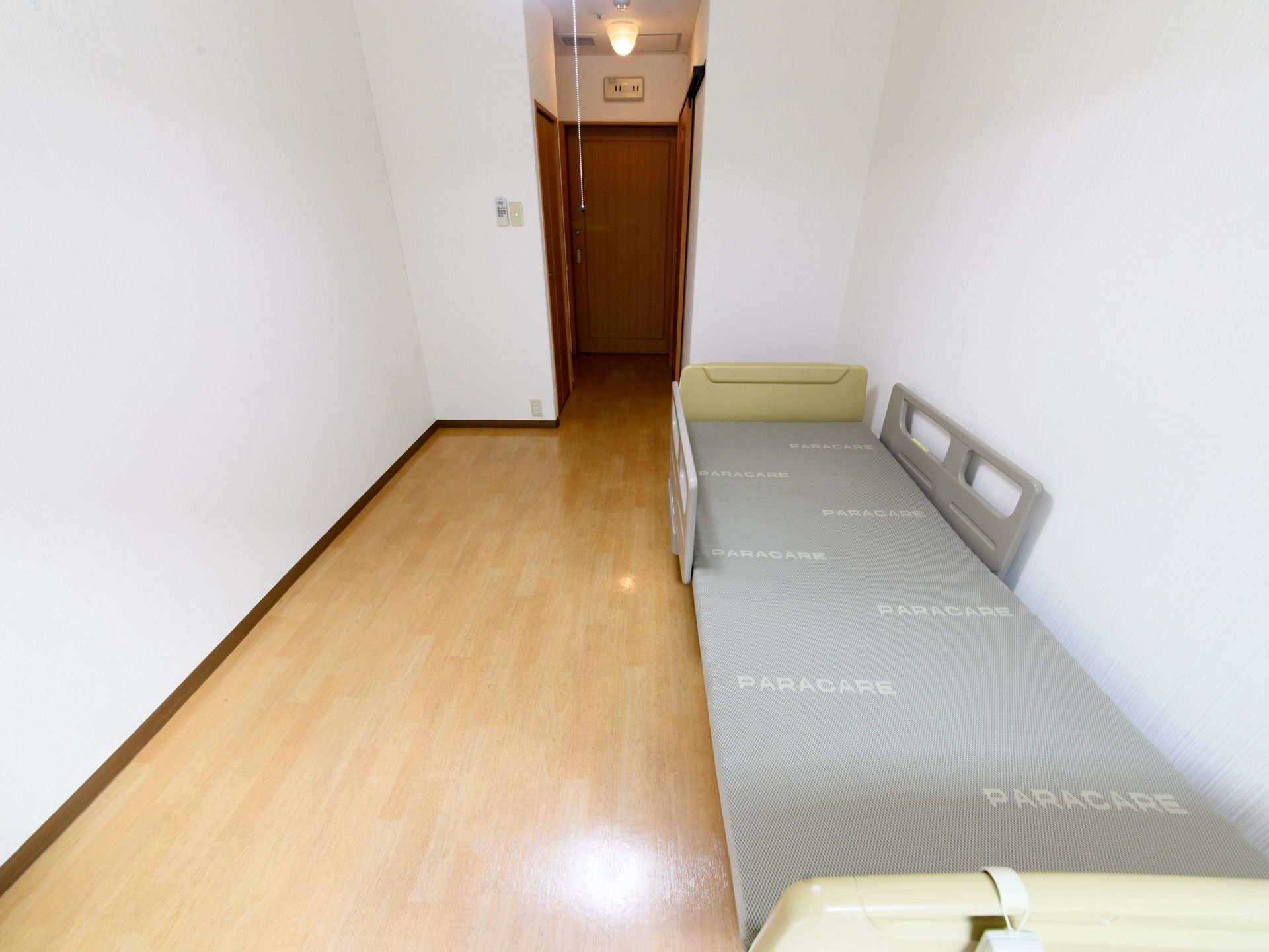 電動ベッドがある。壁は白色で、床は茶色のフローリングである。入口ドアは引き戸式である。壁にエアコンのリモコンが取り付けてある。