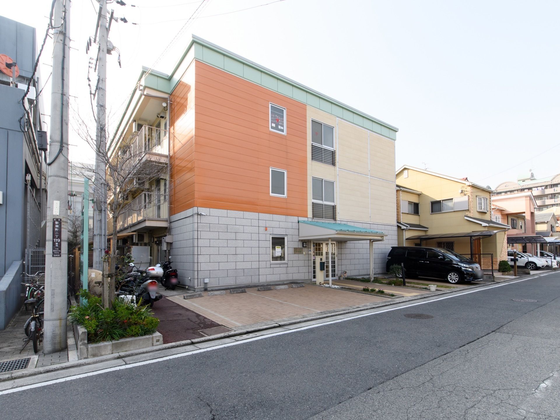 施設は暖色系の色の壁で明るい雰囲気である。玄関の両側に駐車スペースがある。左側にバイクや自転車を置くところもある。