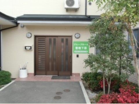 エントランス横には植え込みがあり、赤い花や木が植えられている。茶色い玄関扉の横の壁には施設名の書かれた看板が付けられている。